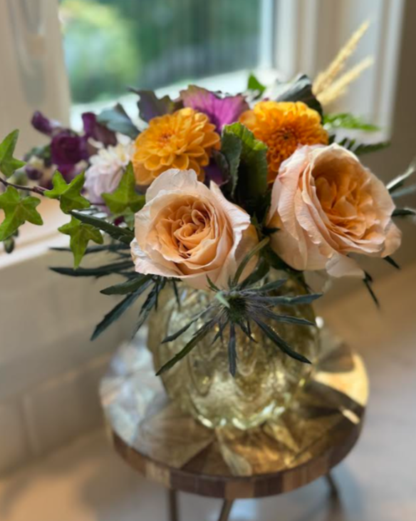 A glass vase with a bouquet of mixed flowers, including roses, marigolds, and other colorful blossoms, sits on a small round wooden table near a window.