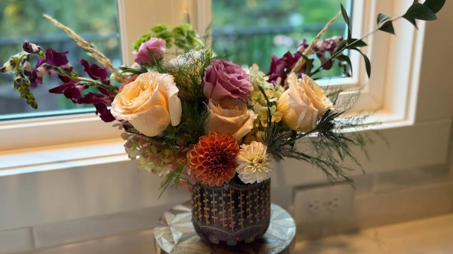 A flower arrangement in a decorative black vase with gold and white patterns, placed on a small wooden stand by a window. The bouquet includes roses, dahlias, snapdragons, and assorted greenery, with a soft natural light illuminating the scene.