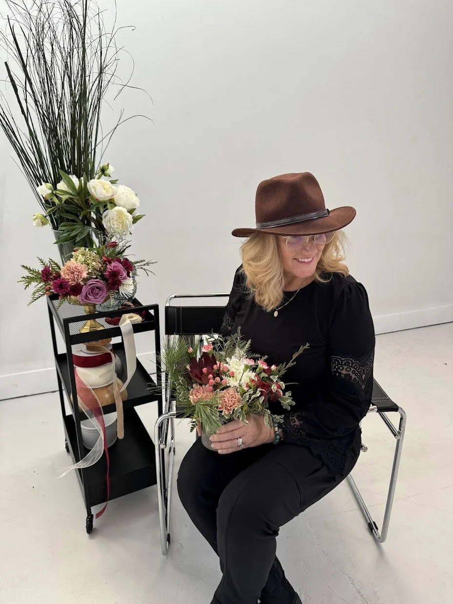 A woman with blonde hair, wearing a brown hat, glasses, a black dress with lace sleeves, and jewelry, is sitting on a black chair holding a bouquet of flowers. Behind her is a flower arrangement with roses, carnations, and greenery on a black cart against a plain white wall.