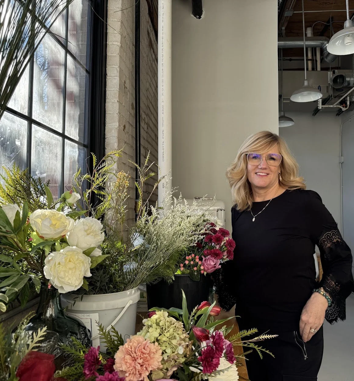 A woman with blonde hair, glasses, wearing a black outfit with lace details, standing next to a table with various flower arrangements in pots and vases.