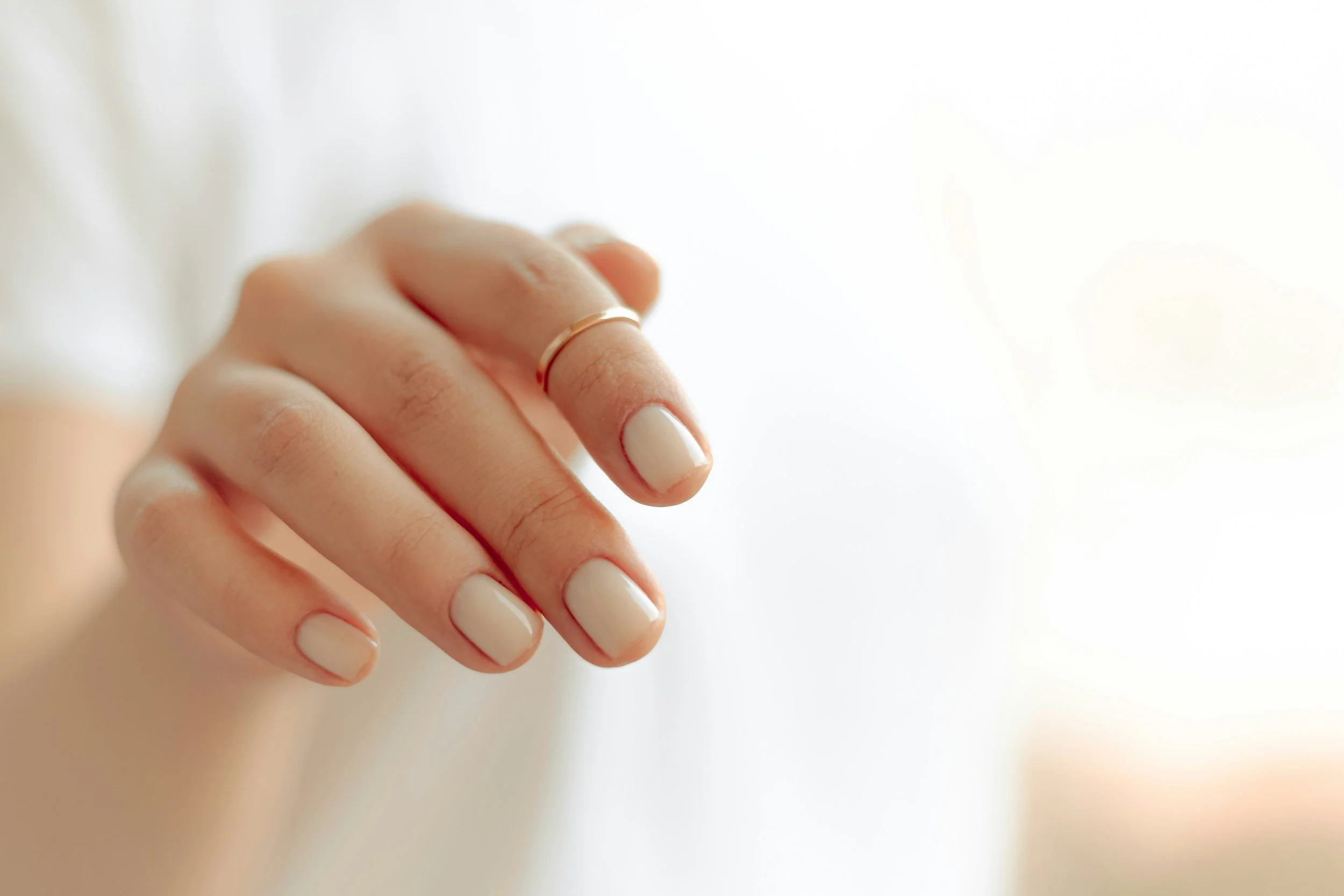 Close-up of a woman's hand with beige painted nails wearing a thin gold ring on her index finger, set against a white background.