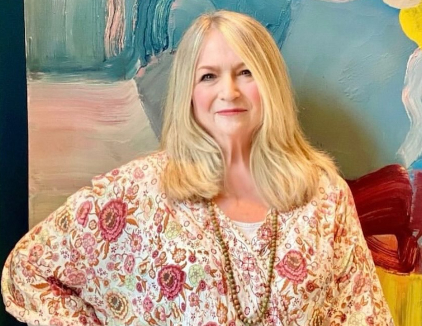 A woman with long blonde hair, wearing a floral patterned blouse and a beaded necklace, standing against an abstract colorful painting background.