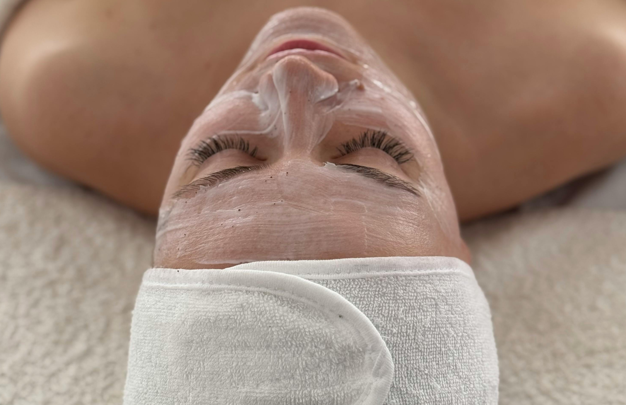 A person with closed eyes lying down, receiving a facial treatment with a white cream or mask, wearing a white towel wrapped around their head.
