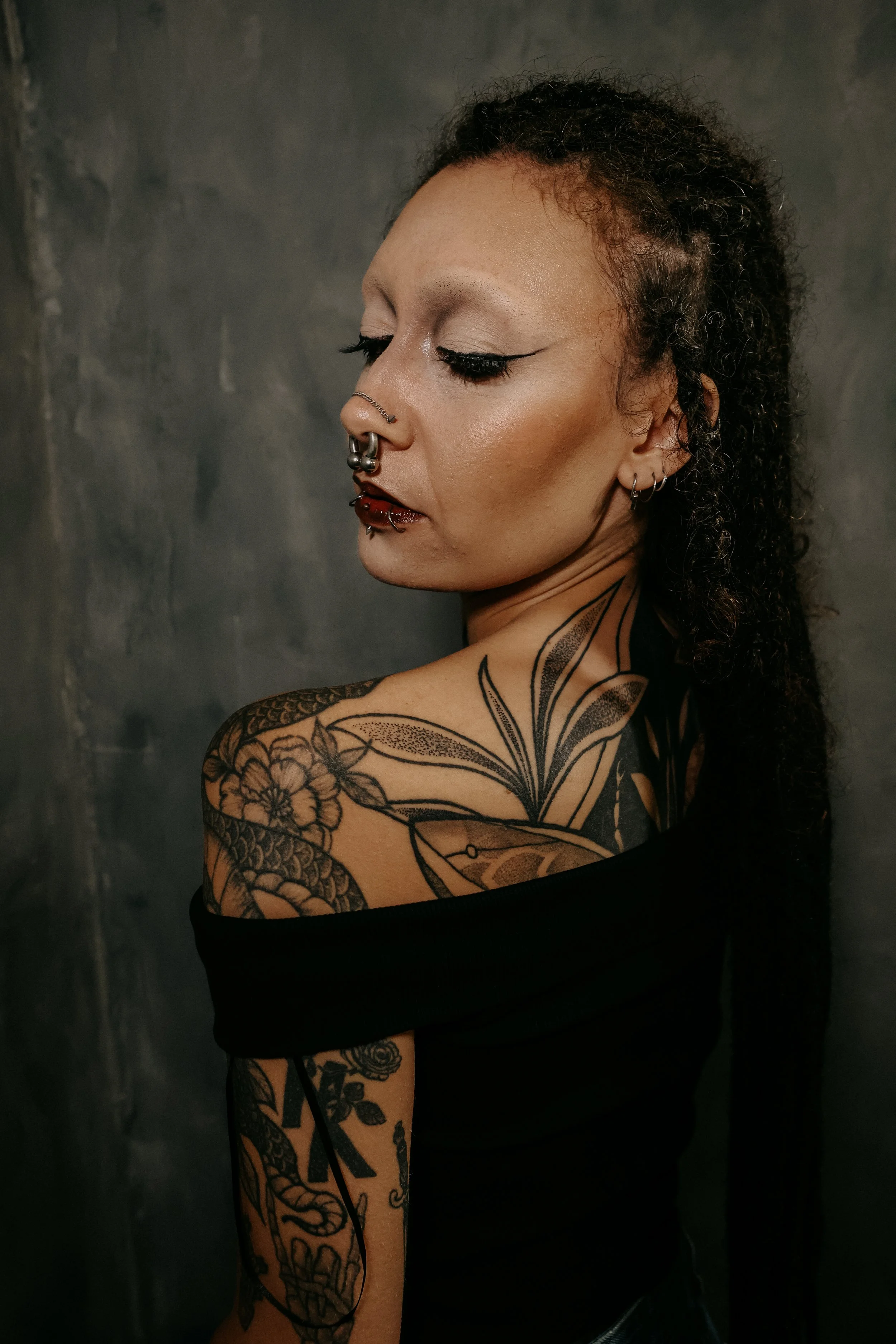A woman with curly hair and piercings, showing detailed tattoos on her shoulder and arm, against a dark background.