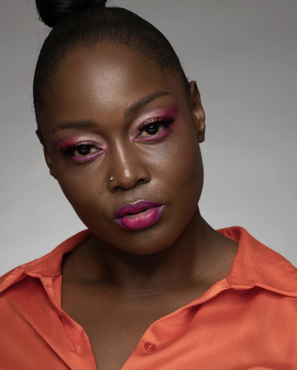 A portrait of a woman with dark skin, wearing bright pink eyeshadow, pink lipstick, and a nose piercing. She has her hair styled in a high bun and is dressed in an orange shirt against a plain gray background.