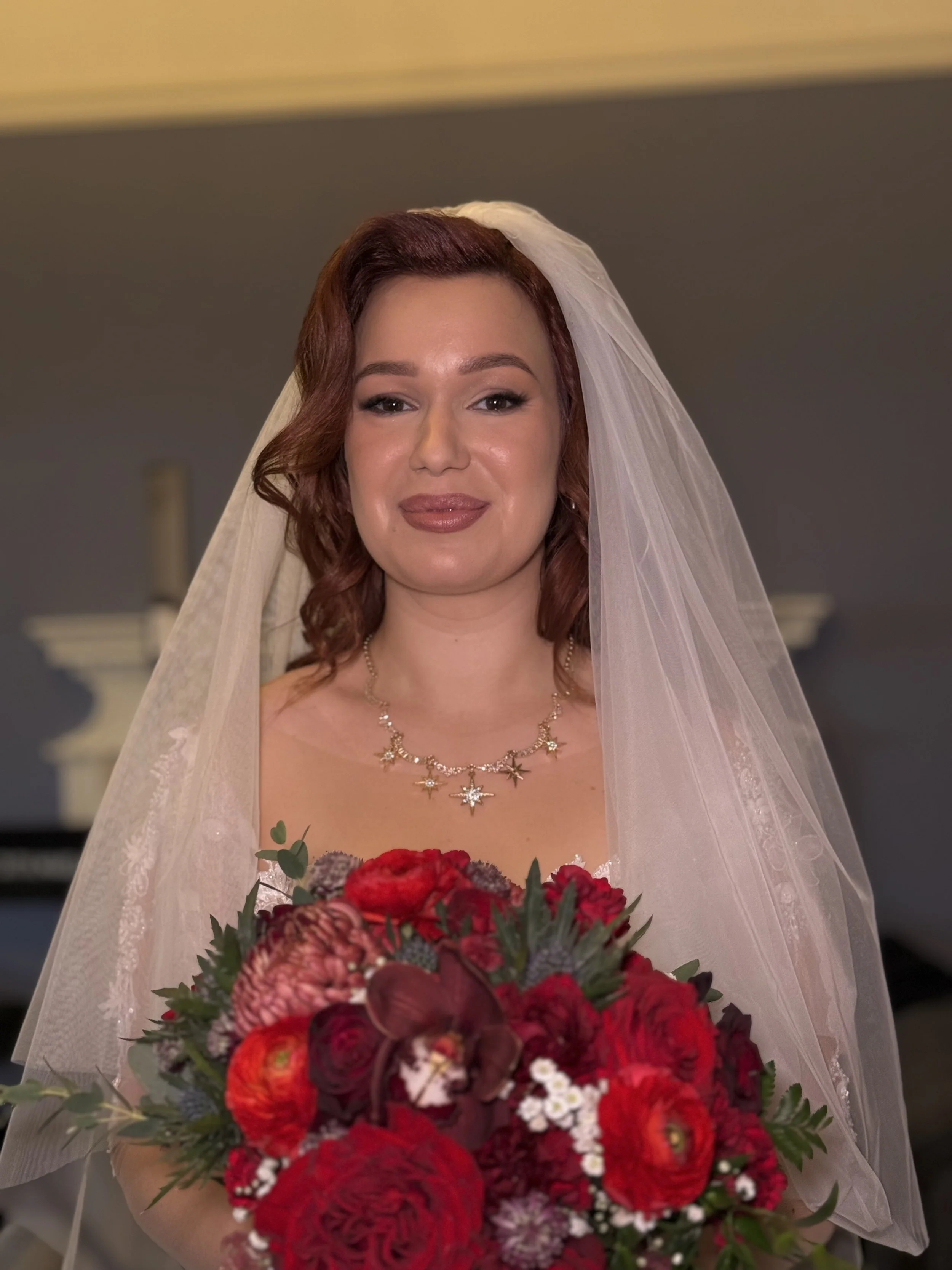A bride with auburn hair, wearing a white lace veil and a gold star-shaped necklace, holding a bouquet of red and burgundy flowers.