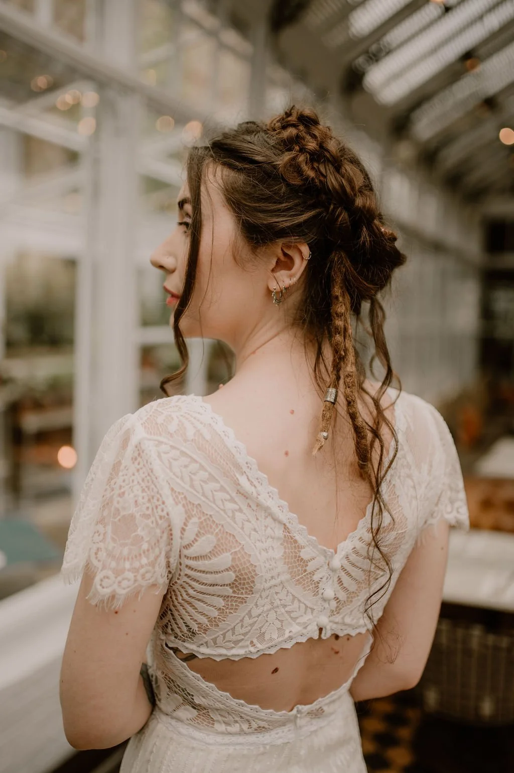 A woman with braided and loose curly hair, wearing a white lace top with buttons, stands indoors with her back to the camera, in a room with glass windows and blurred background lighting.