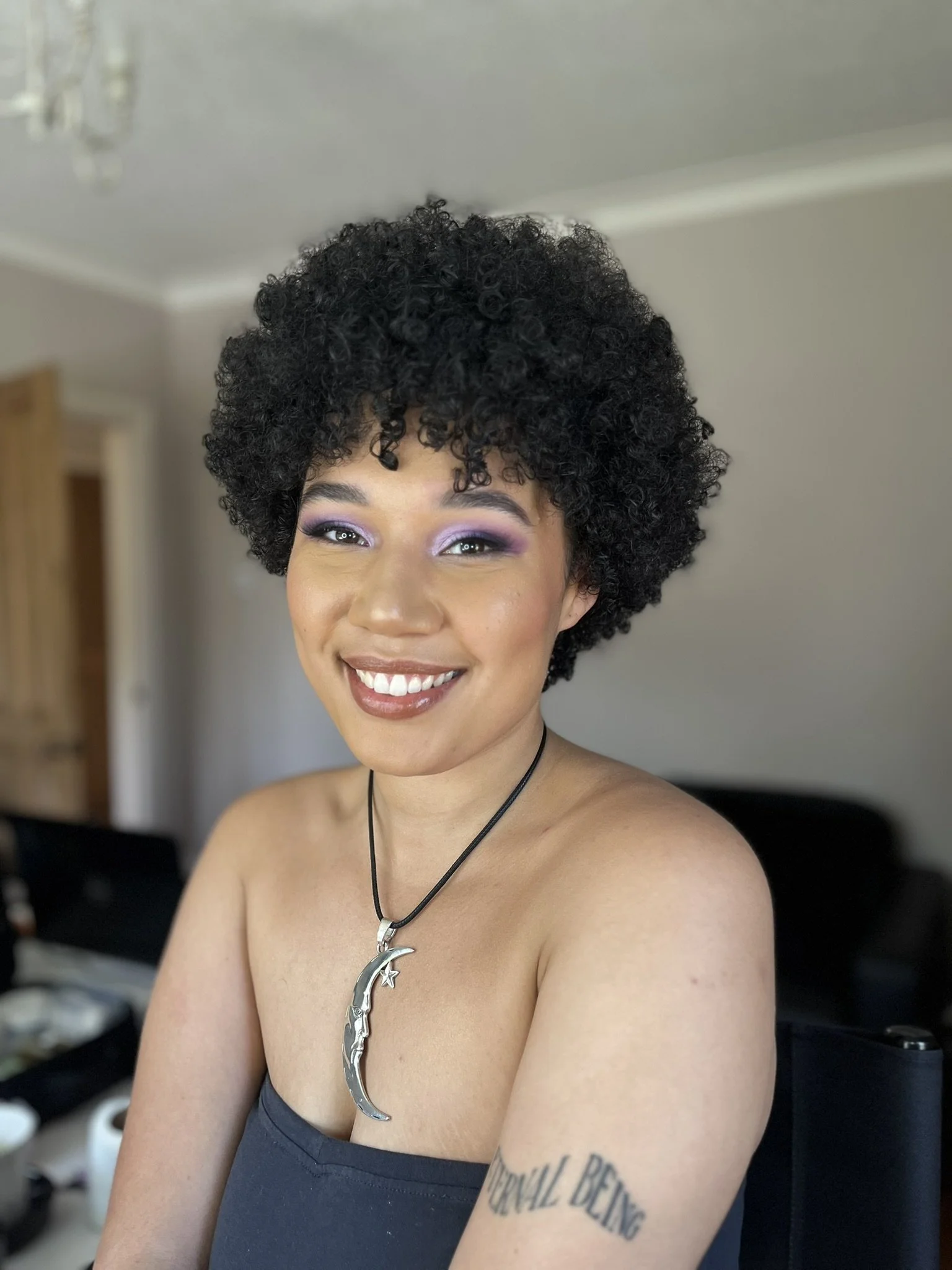 A woman with dark, curly hair smiling and wearing purple eyeshadow, a black top, and a crescent moon necklace, sitting indoors.