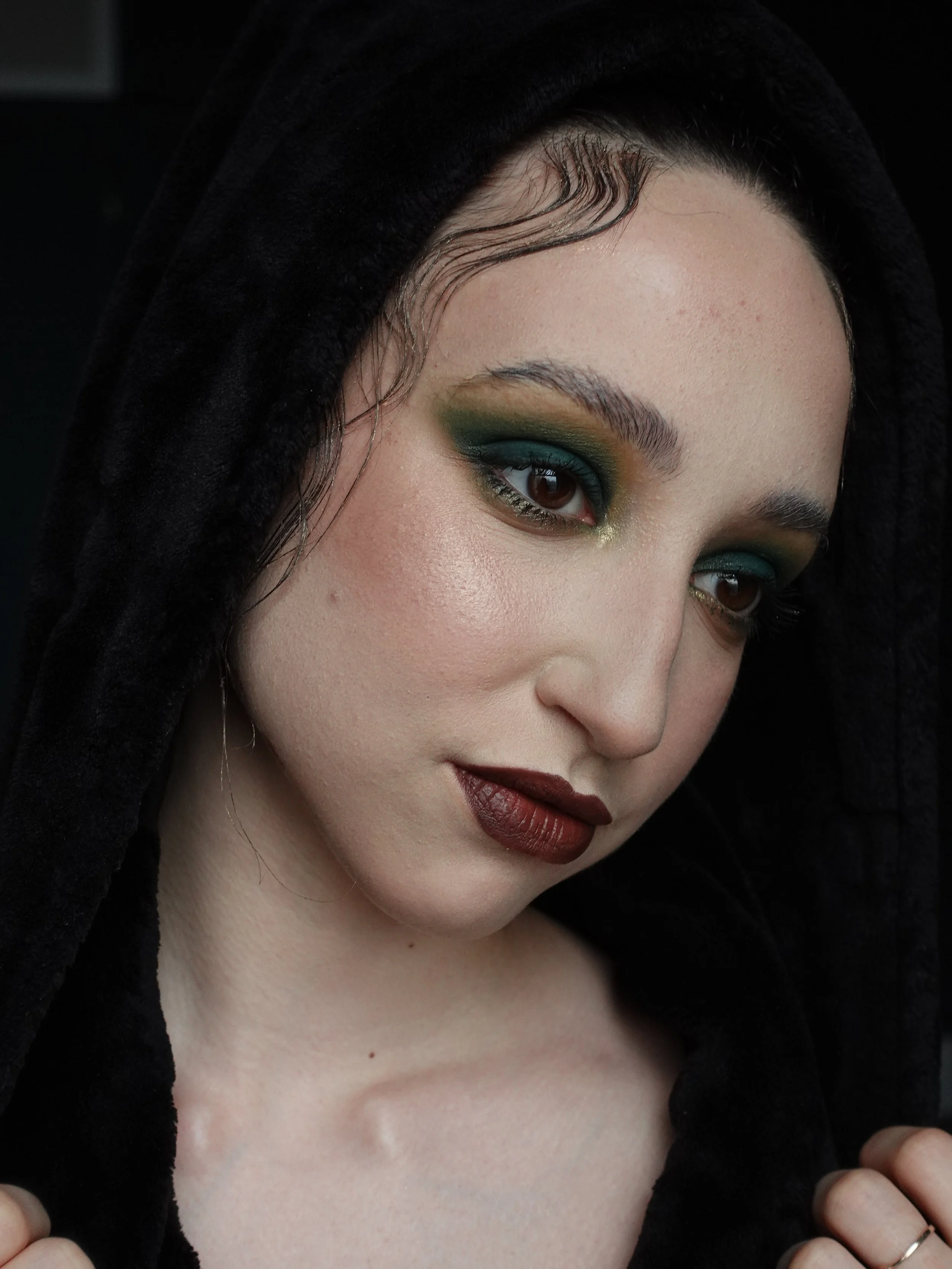 A woman with dramatic makeup, including green eyeshadow, dark red lipstick, and well-defined eyebrows, wearing a black hooded garment.