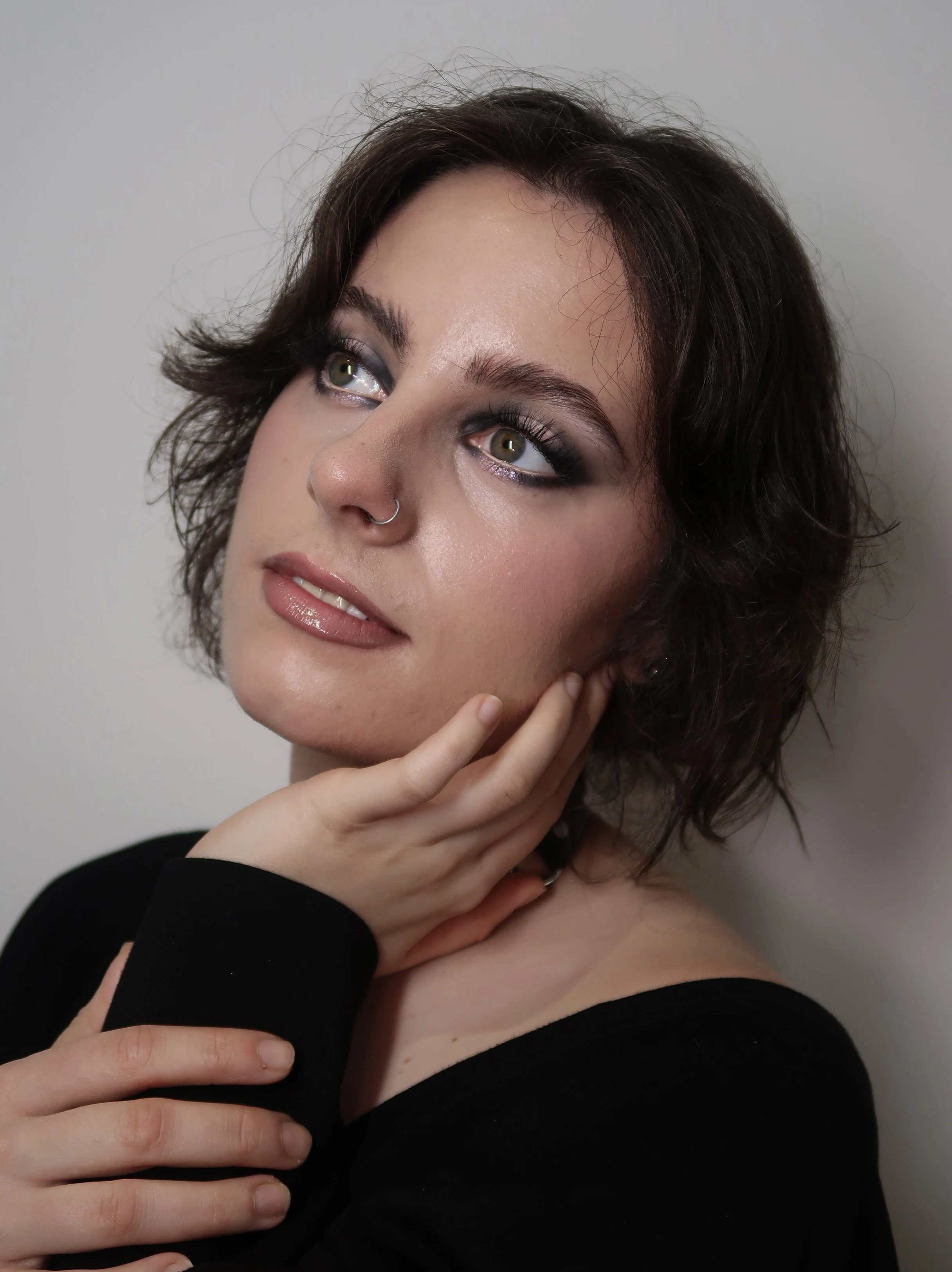 A young woman with short, wavy brown hair, light skin, and hazel eyes. She is touching her face with her right hand, with her left hand resting on her right shoulder. She is wearing a black top, subtle makeup, and a nose ring.