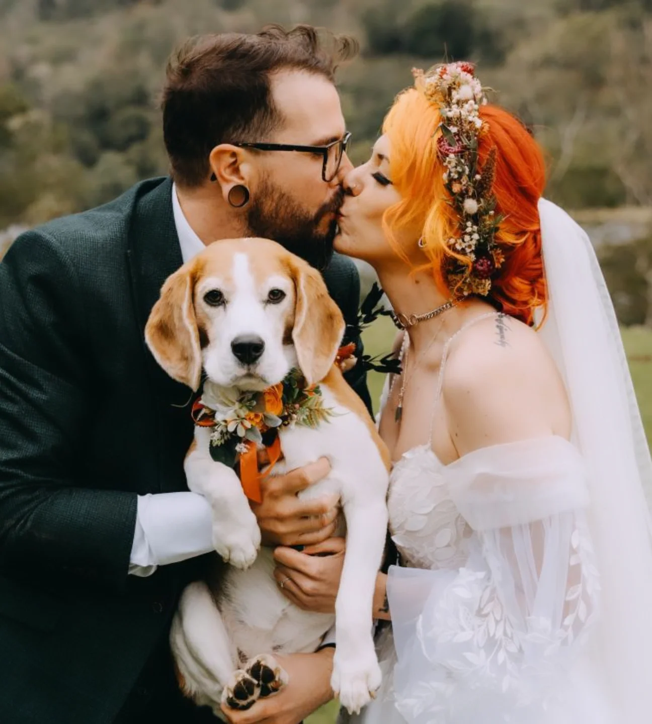 A couple in wedding attire kiss while holding a beagle dog decorated with flowers near a lake or pond outdoors.