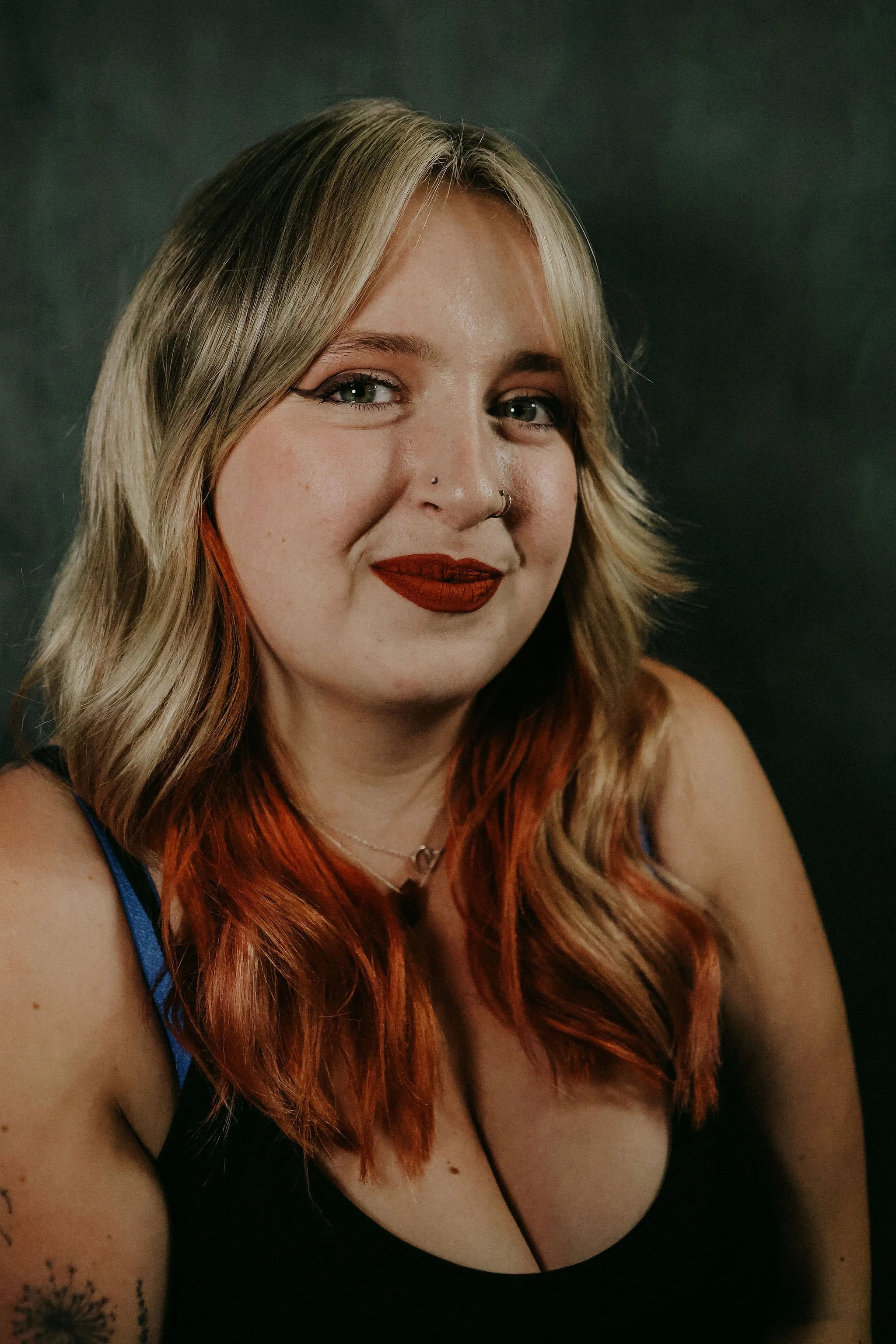 Close-up portrait of a young woman with blonde hair and red tips, wearing red lipstick, a black top with blue straps, and a silver necklace, smiling slightly against a dark background.