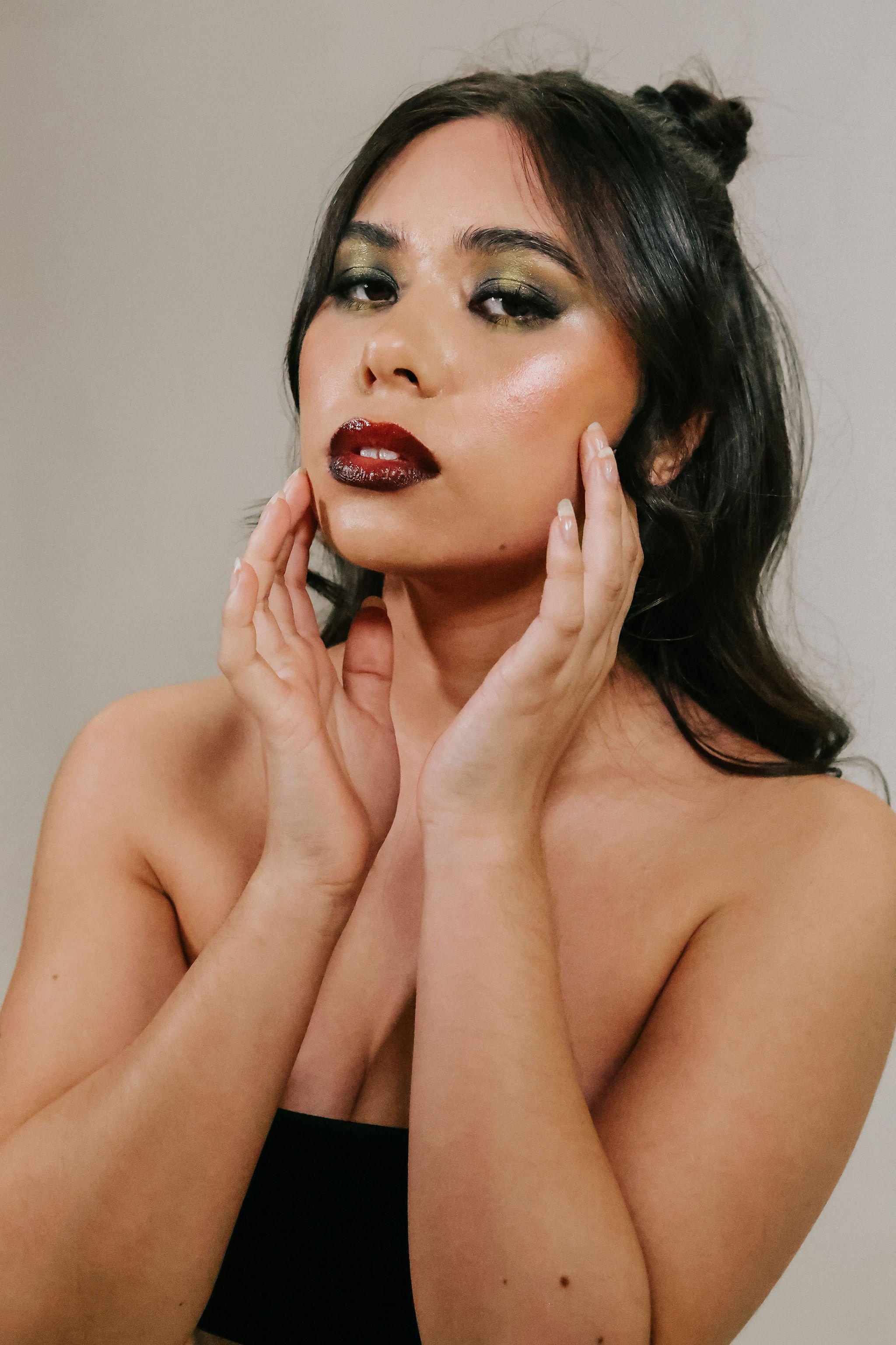 A woman with dark hair styled in loose waves, wearing bold makeup including dark red lipstick and gold eyeshadow, gently touching her face with both hands, wearing a black strapless top, against a plain background.