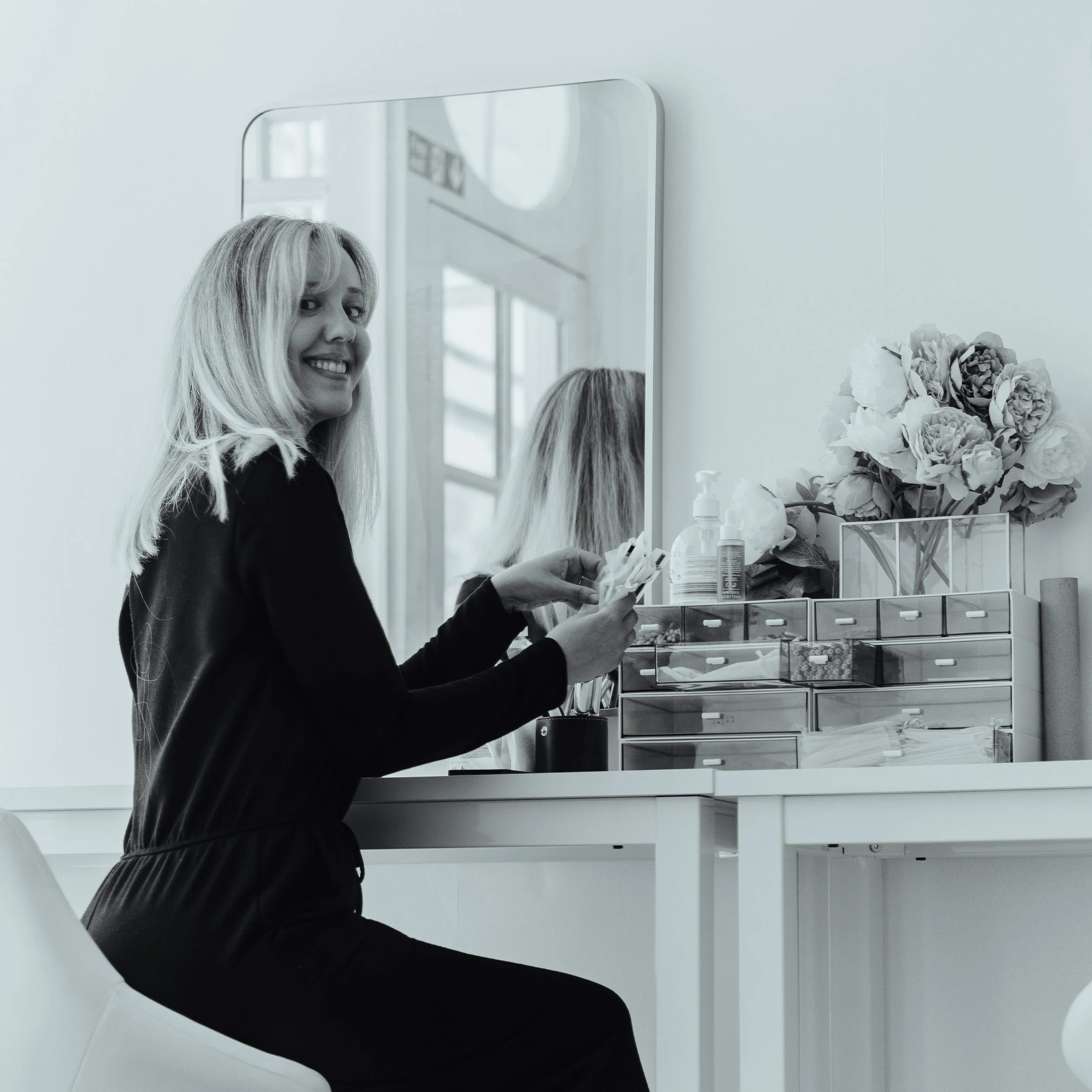 Woman sitting at a makeup vanity, smiling, with a mirror, makeup supplies, and a flower arrangement.