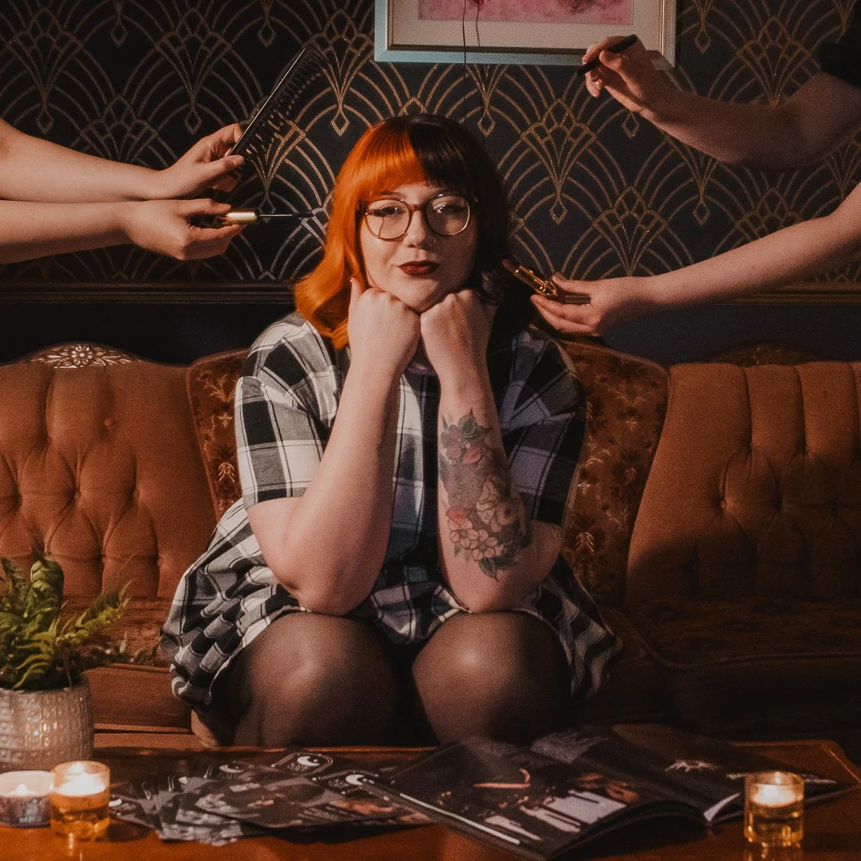 A woman with red and black hair, glasses, and tattoos sits on a vintage sofa, resting her chin on her fists, with multiple people applying makeup and holding brushes around her in a dimly lit room with dark patterned wallpaper.