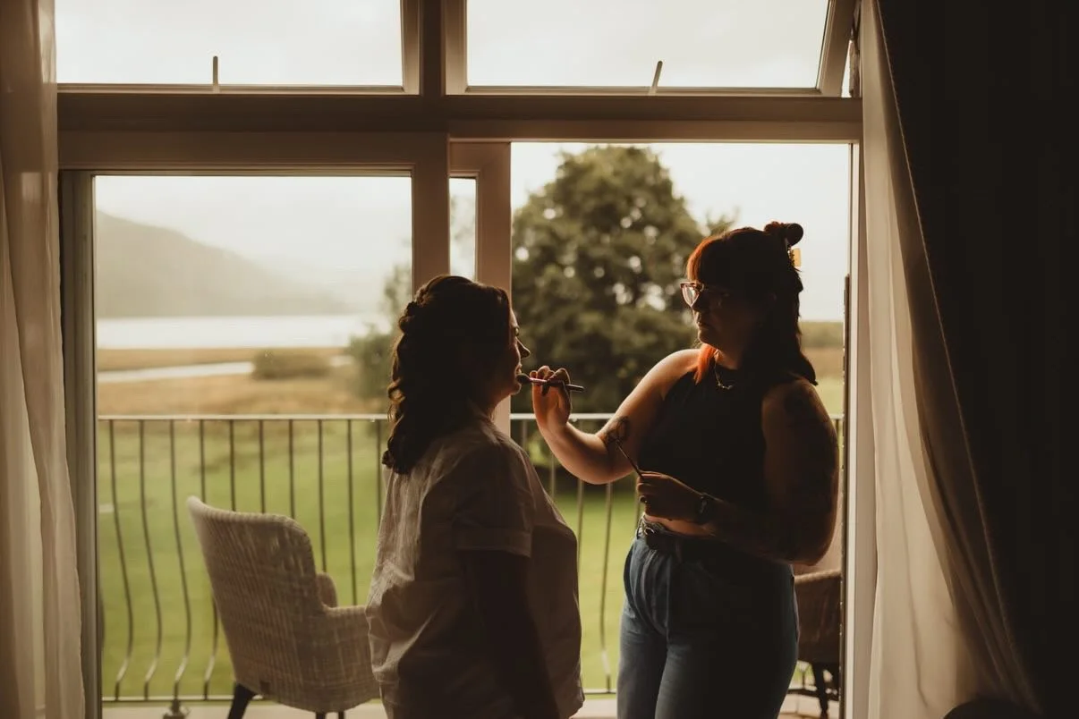 A makeup artist applying makeup to a woman sitting near a large window with a scenic view of trees and a body of water.