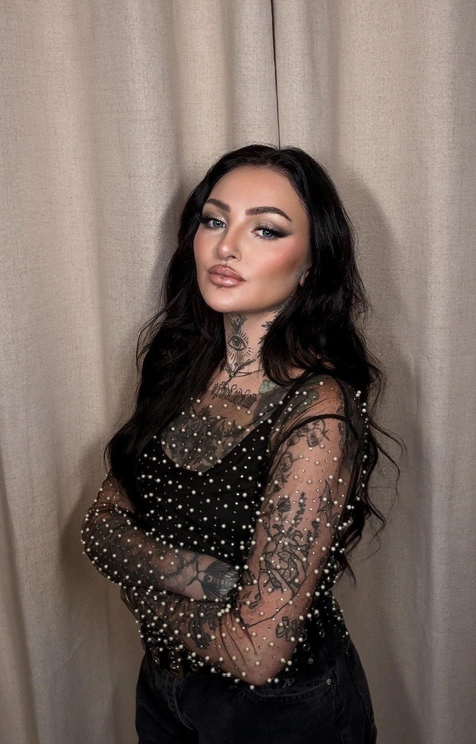 A woman with long black hair and tattoos wearing a black dress with pearl embellishments, standing in front of a beige curtain.