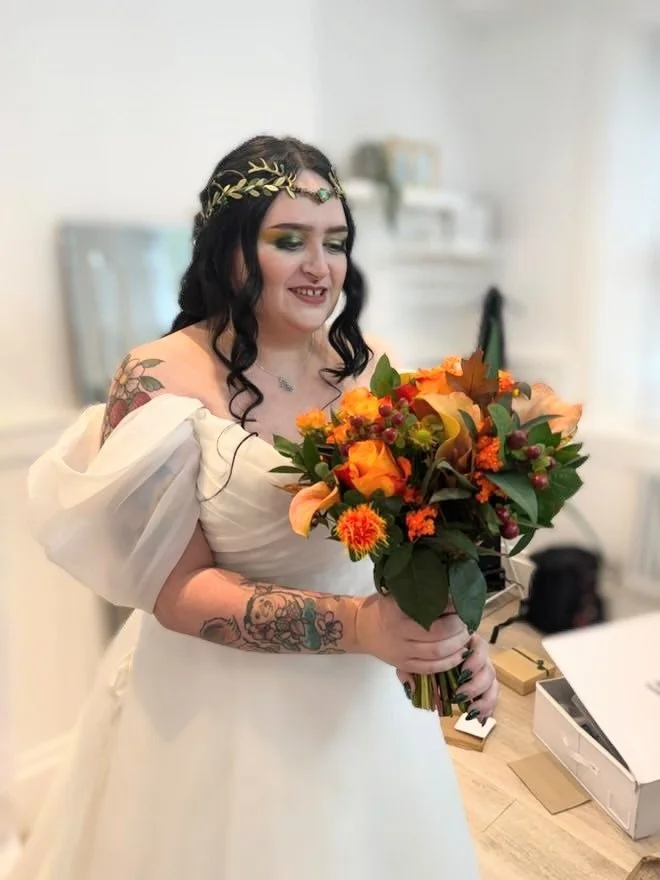 A bride with dark wavy hair, makeup, and tattoos on her arms, wearing an off-shoulder white wedding dress and a gold leaf crown, holding a bouquet of orange and yellow flowers.