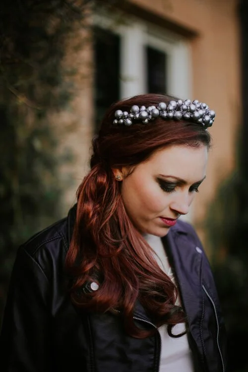 Woman with red hair wearing a pearl and silver headband, looking down, outdoors with a blurred building and greenery background.