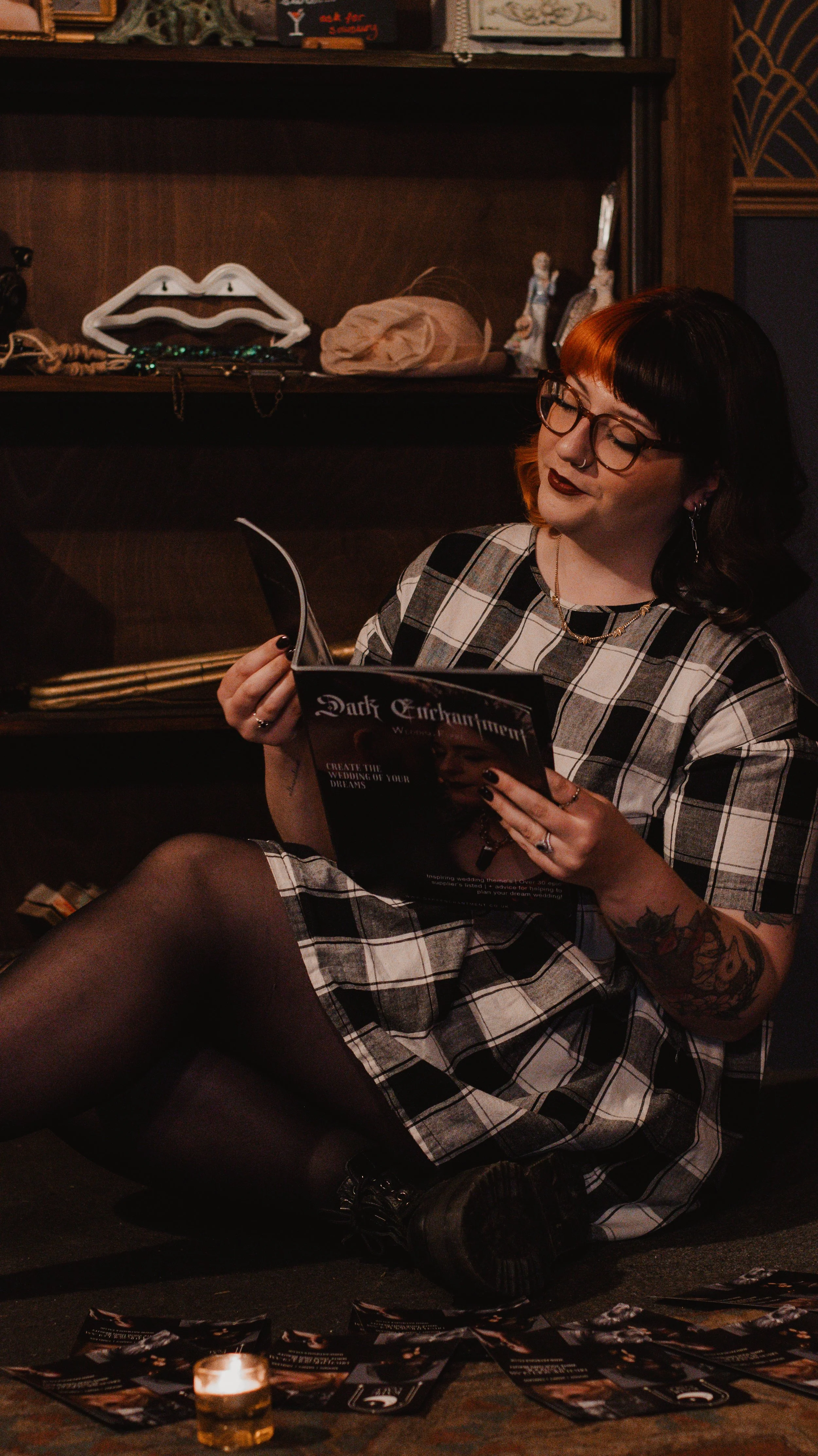 A woman with black and red hair, glasses, and tattoos sitting on the floor, reading a magazine titled 'Dark Enchantment' in a dimly lit room, with a candle and scattered papers on the floor.