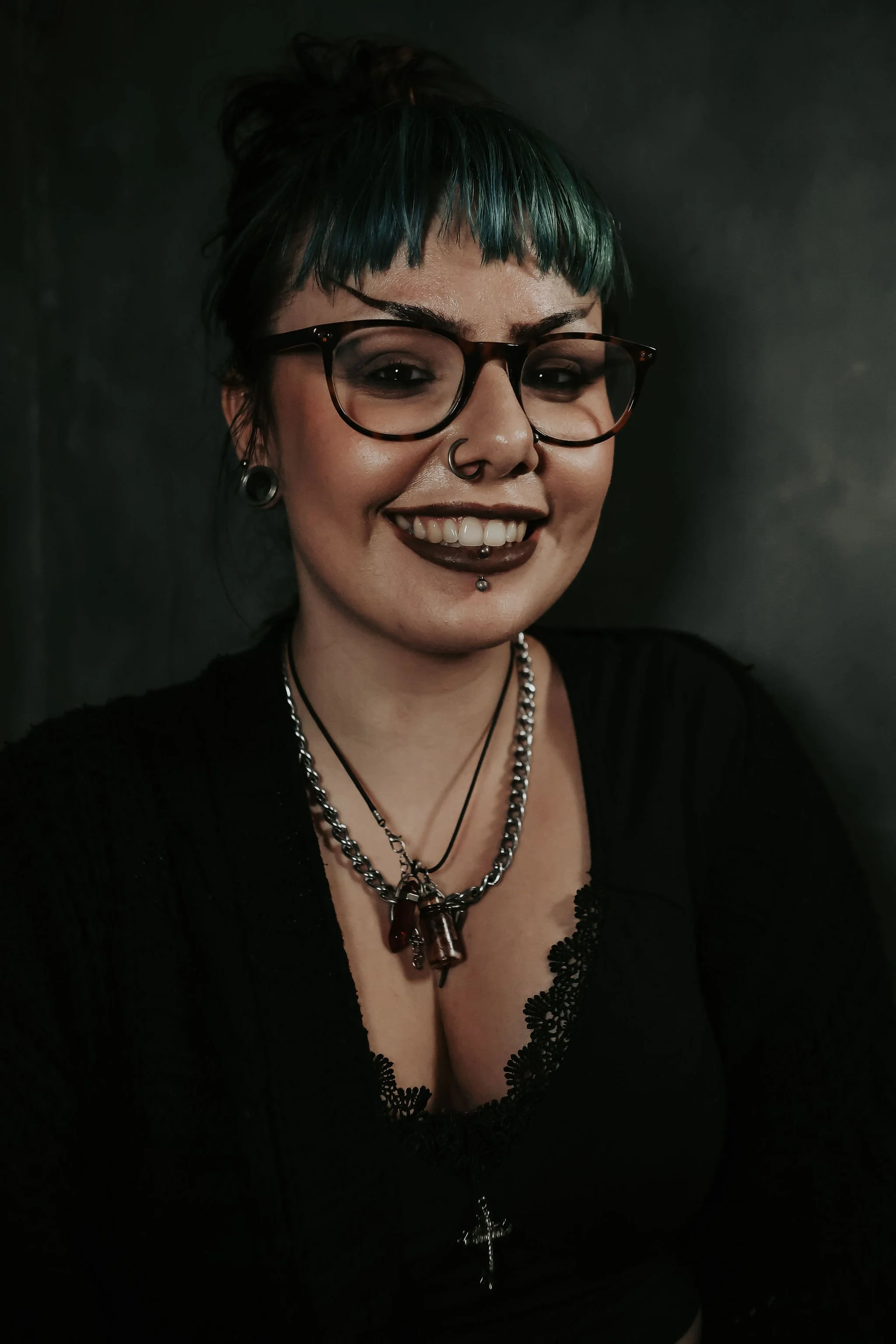 Young woman with short blue and green hair, wearing glasses, multiple piercings, necklaces, and a black top with lace detail, smiling against a dark background.