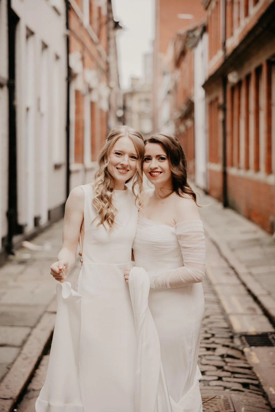 Two women wearing wedding dresses standing on a cobblestone alleyway with brick buildings on each side.