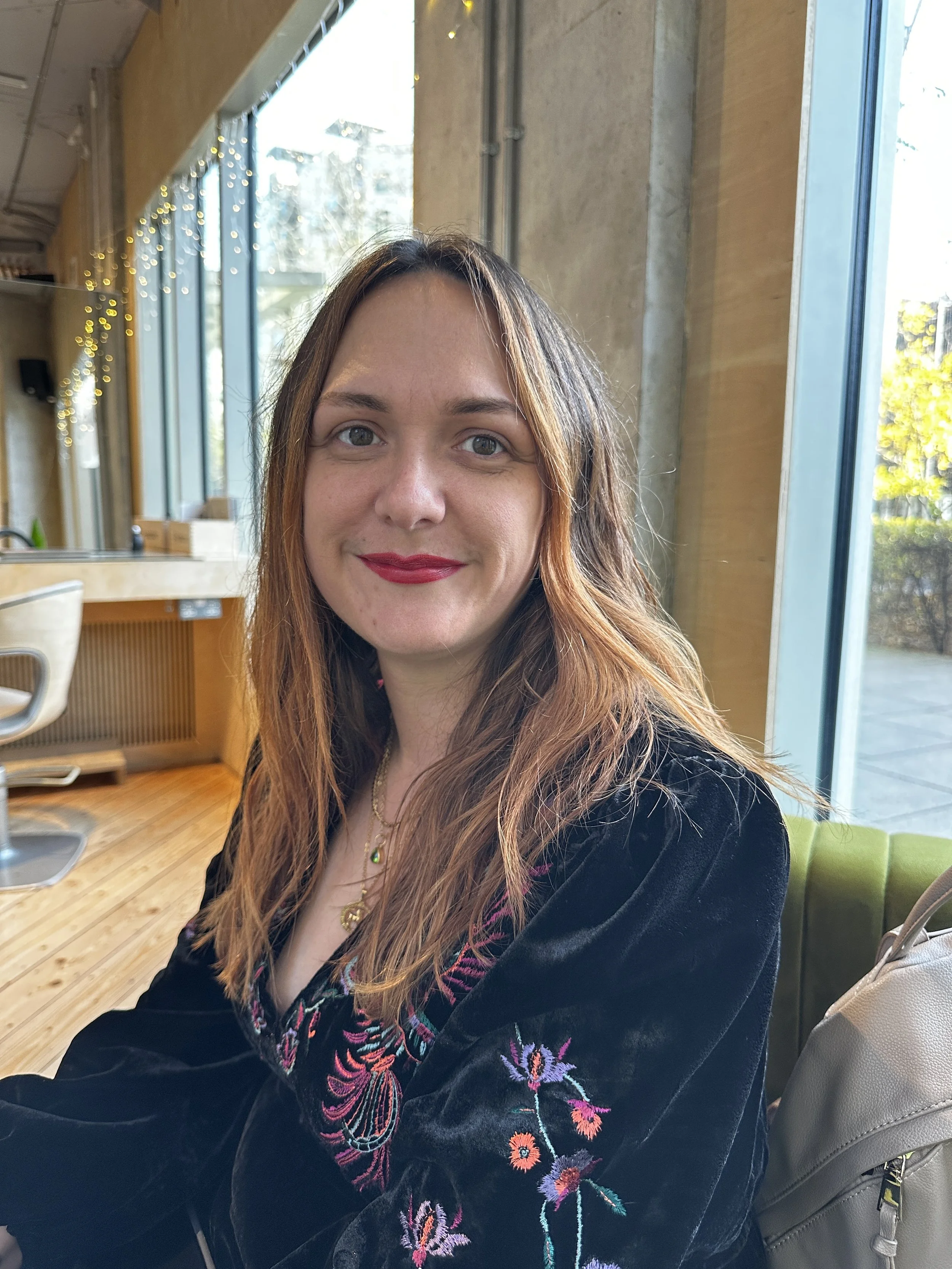 A woman with long reddish-brown hair, wearing red lipstick and a black embroidered jacket, sitting indoors near large windows with sunlight, in a room with wooden flooring and wall decor.