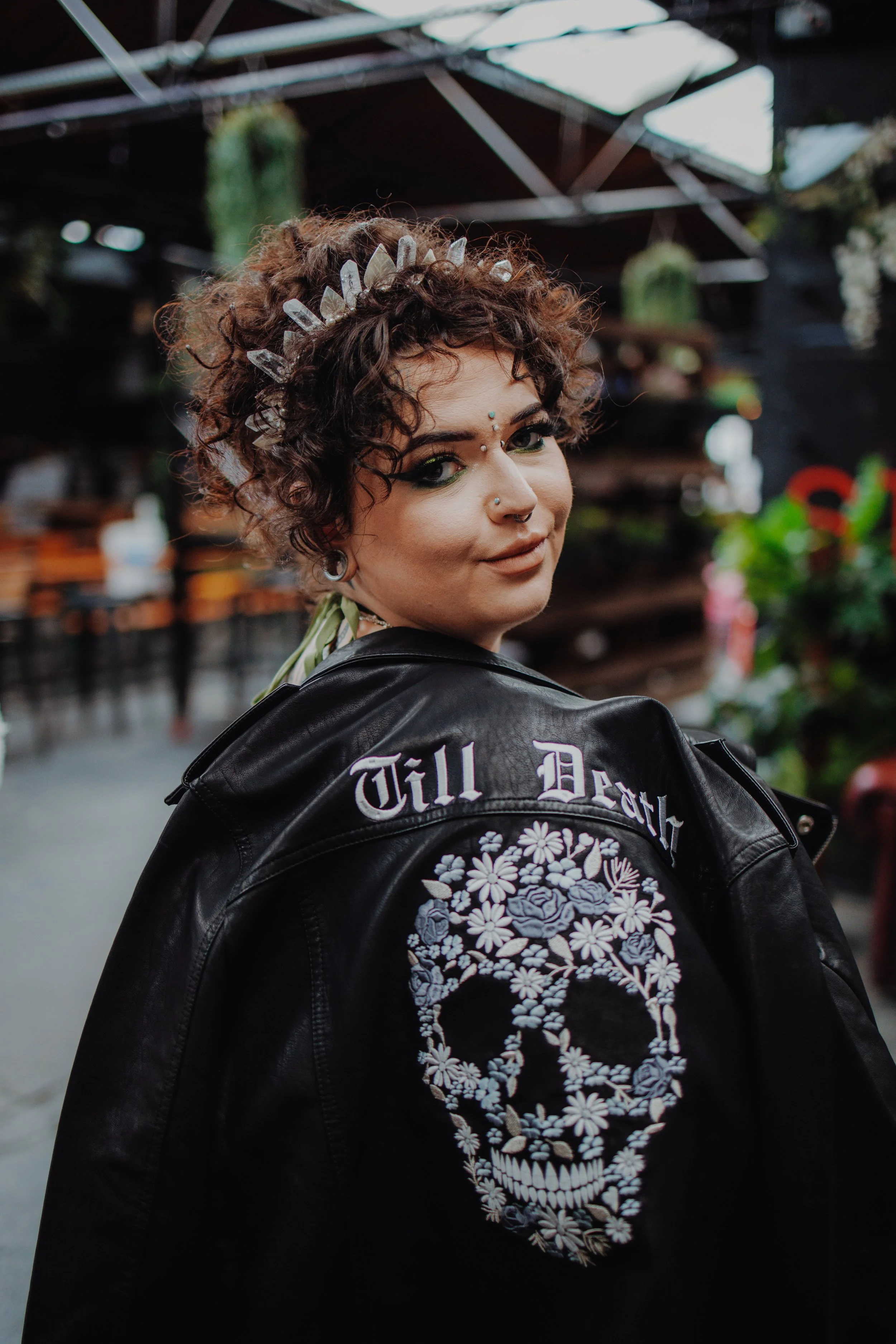 A woman with curly hair wears a black leather jacket with a skull design and the words 'Till Death' and a crystal tiara, standing in an indoor space with plants and industrial decor.