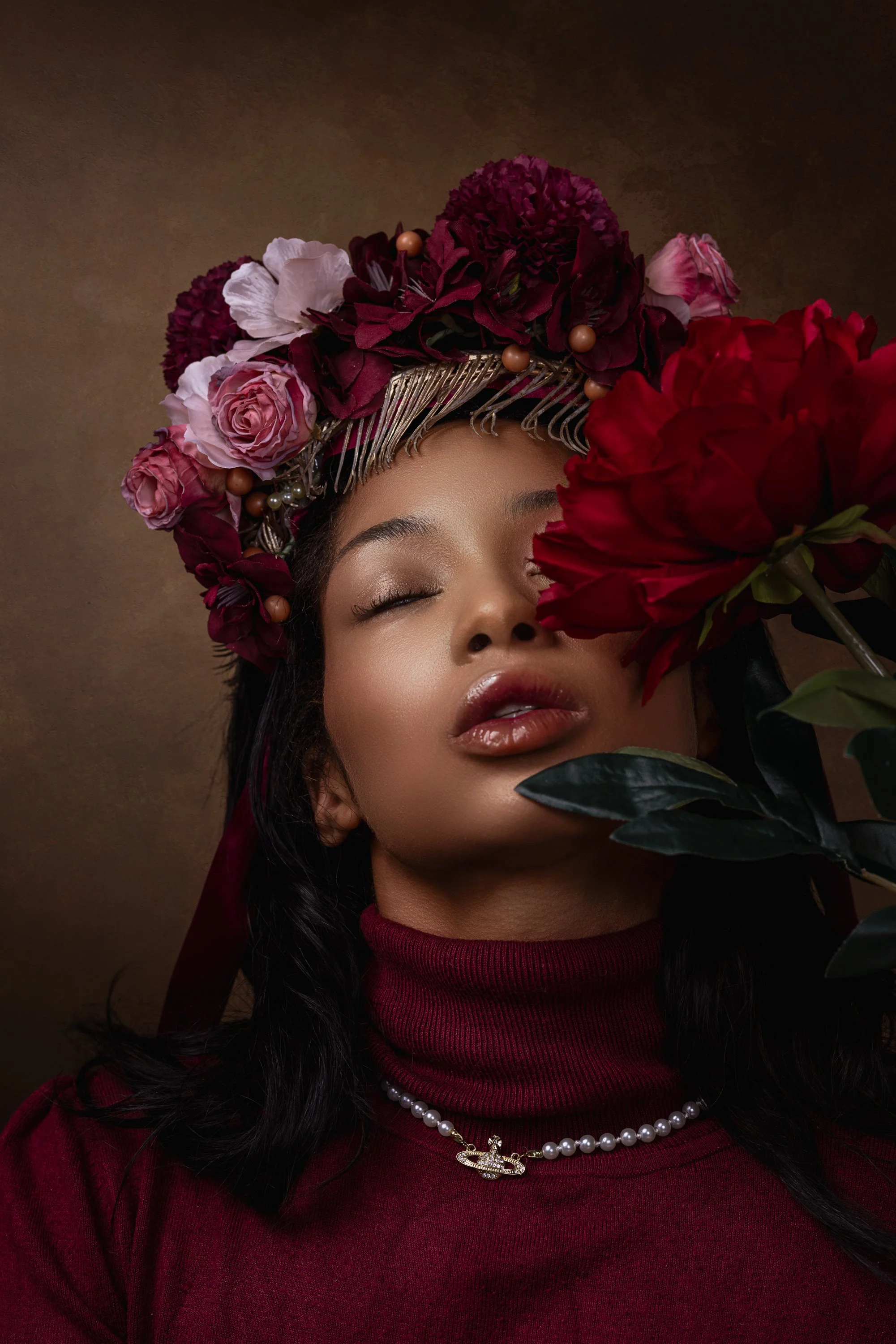 A woman with medium skin tone, wearing a floral crown made of pink, purple, and deep red flowers, a pearl necklace with a pendant, and a red turtleneck, poses with her eyes closed in front of a brown background, partially obscured by large red flower