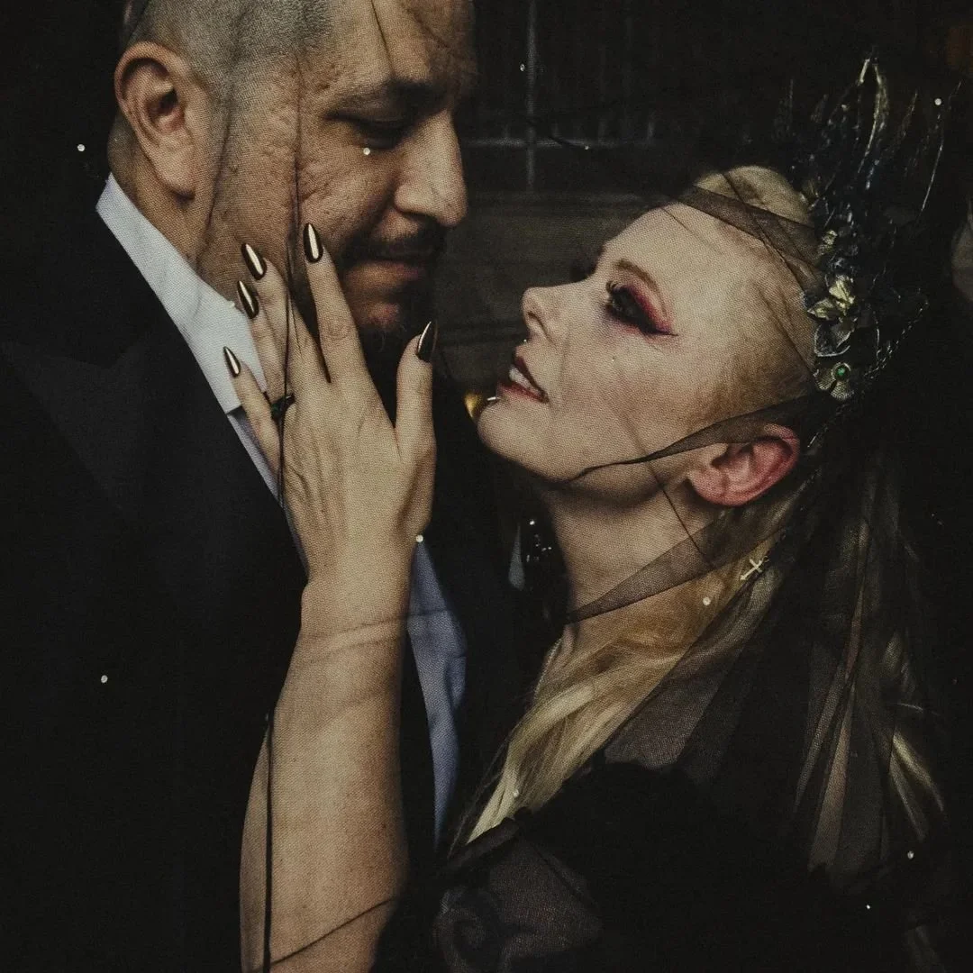 A man in a tuxedo and a woman dressed as a witch or fantasy character are close together, sharing an intimate moment with their foreheads nearly touching.