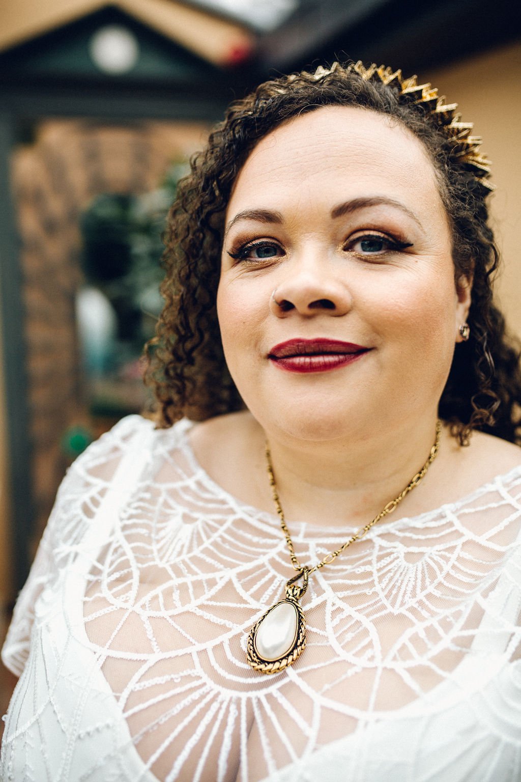 Close-up of a woman with curly hair wearing a white lace dress, gold necklace with large pendant, and red lipstick, smiling at the camera.