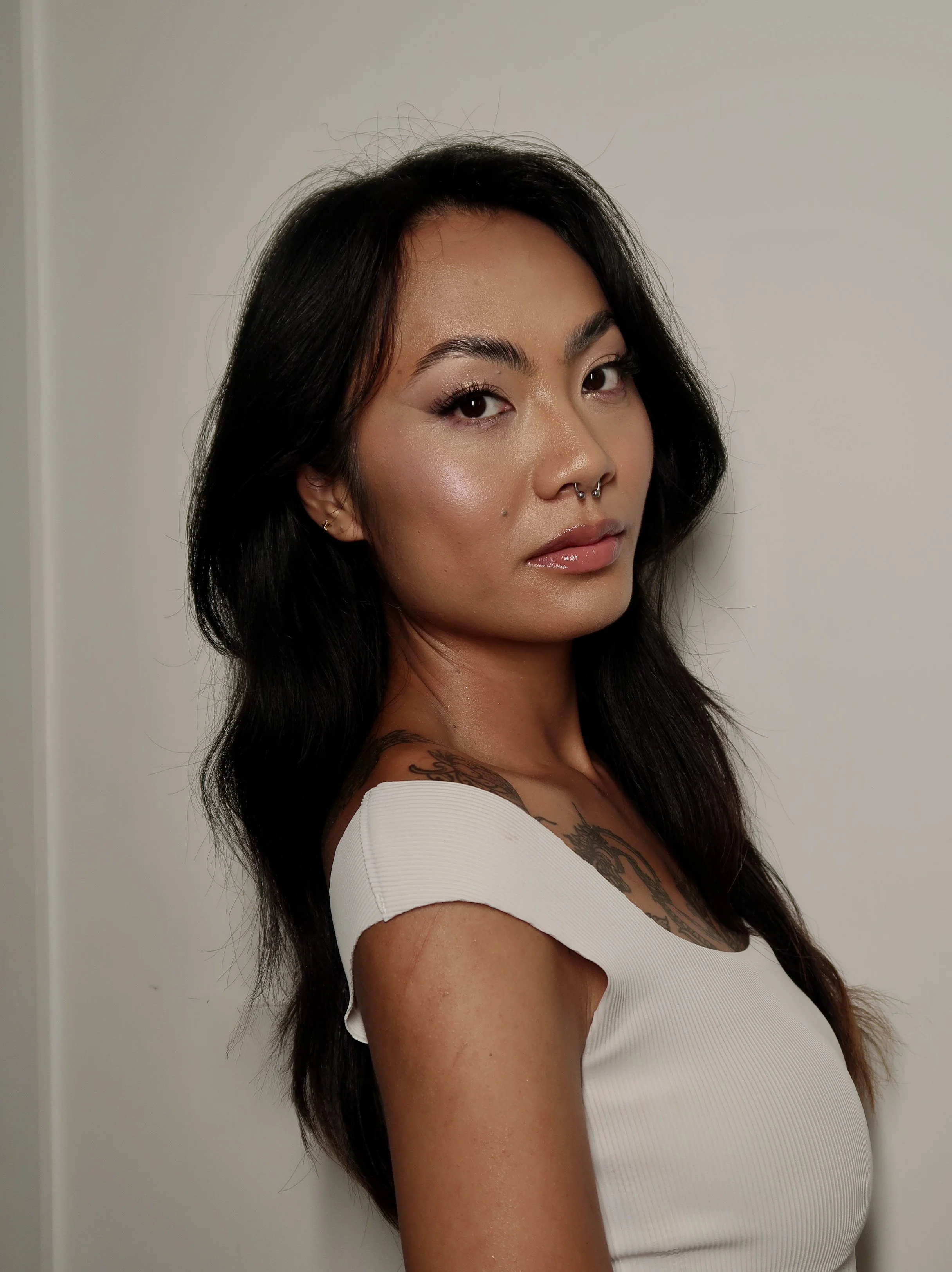 Portrait of a woman with long dark hair, wearing a white top, and a tattoo on her chest, standing against a plain white background.