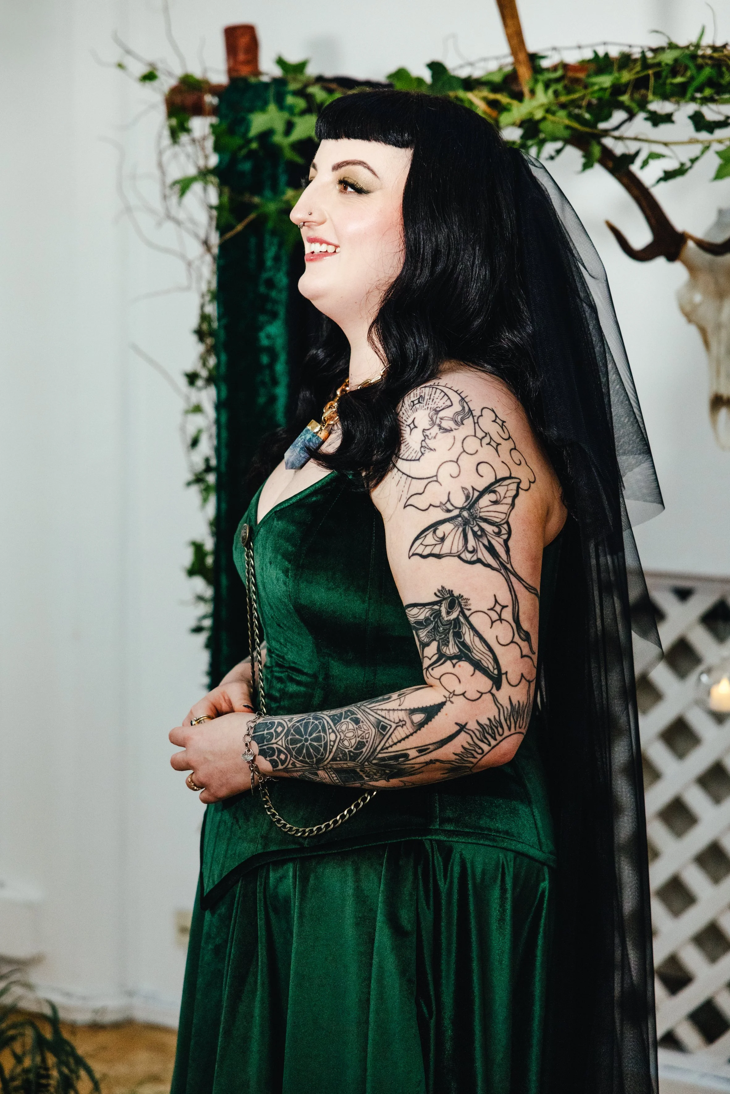 A woman with black hair, tattoos on her arm and shoulder, wearing a green dress and black veil, smiling and standing indoors.