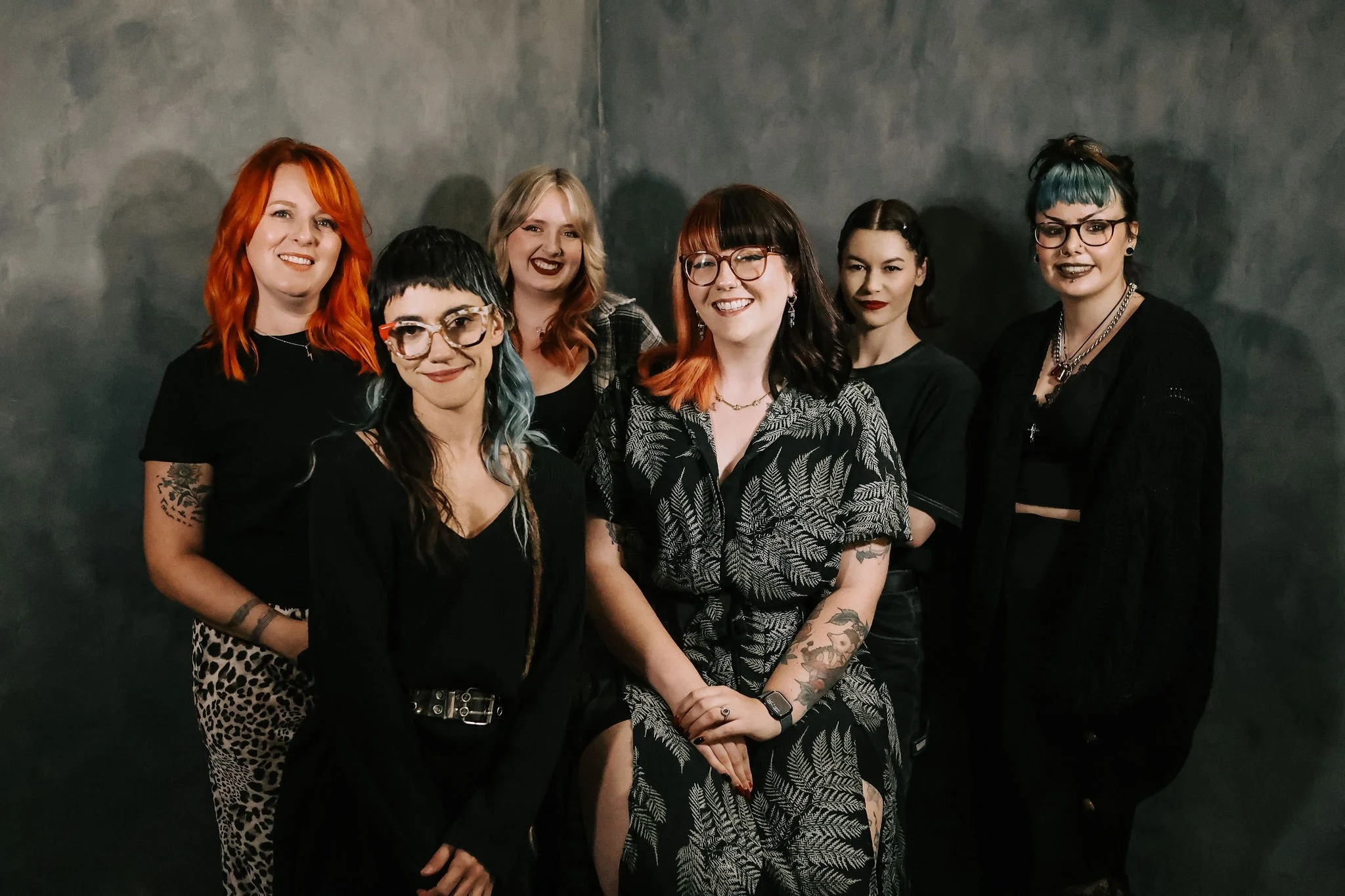 Group of seven women with colorful hair and tattoos, dressed in black, standing against a dark gray wall.