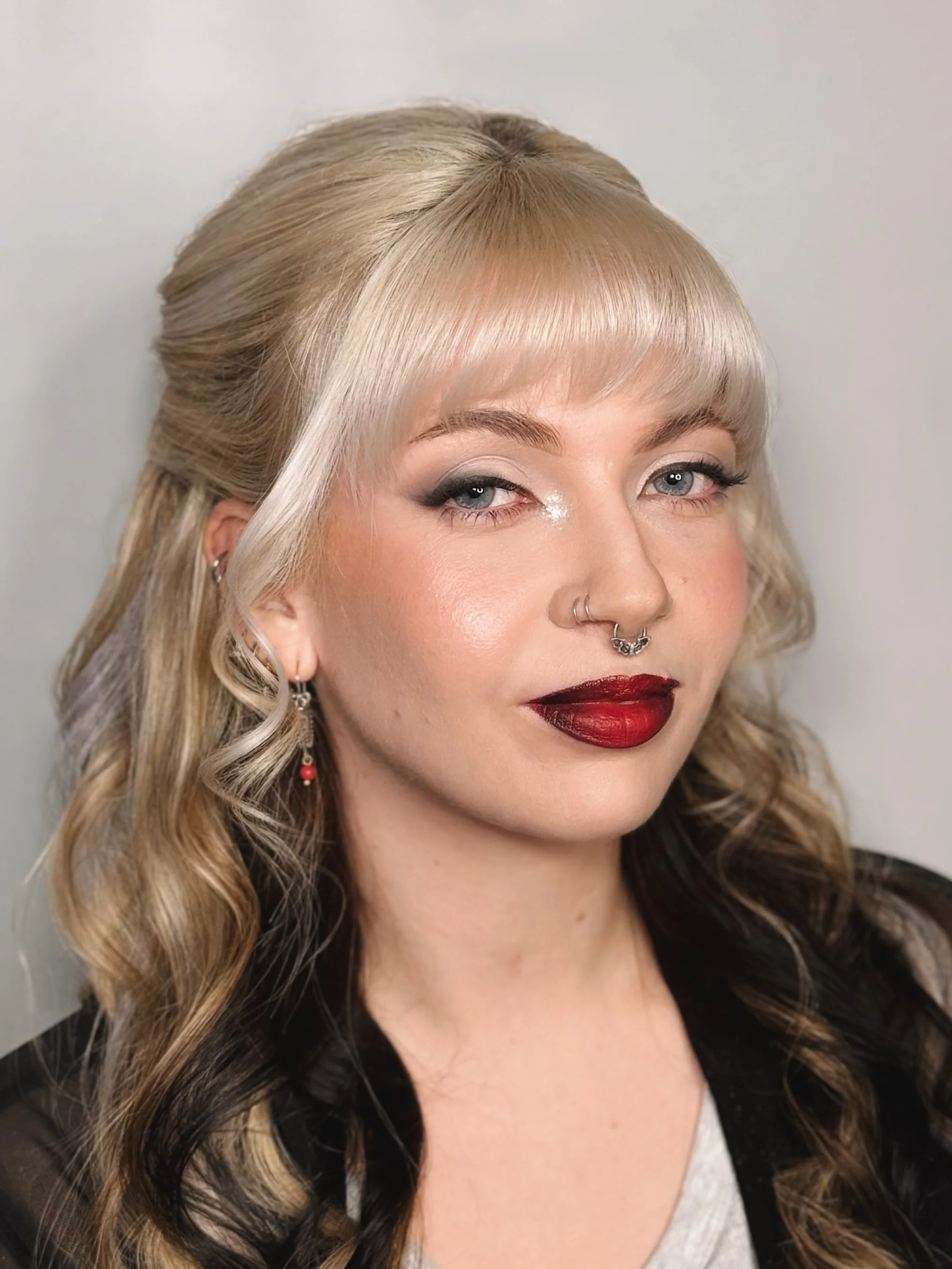 A woman with blonde and black wavy hair, blue eyes, and makeup including dark eyeshadow and red lipstick, wearing earrings and a septum ring, looking at the camera with a slight smile.
