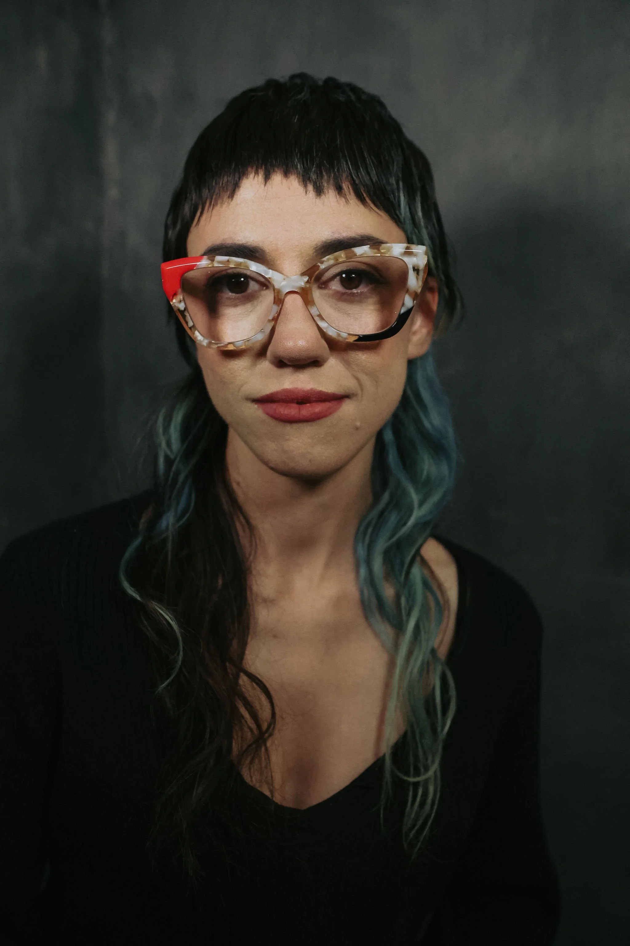 A woman with short black hair and blue-tinted wavy hair wearing large, patterned eyeglasses and a black top, standing against a dark background.