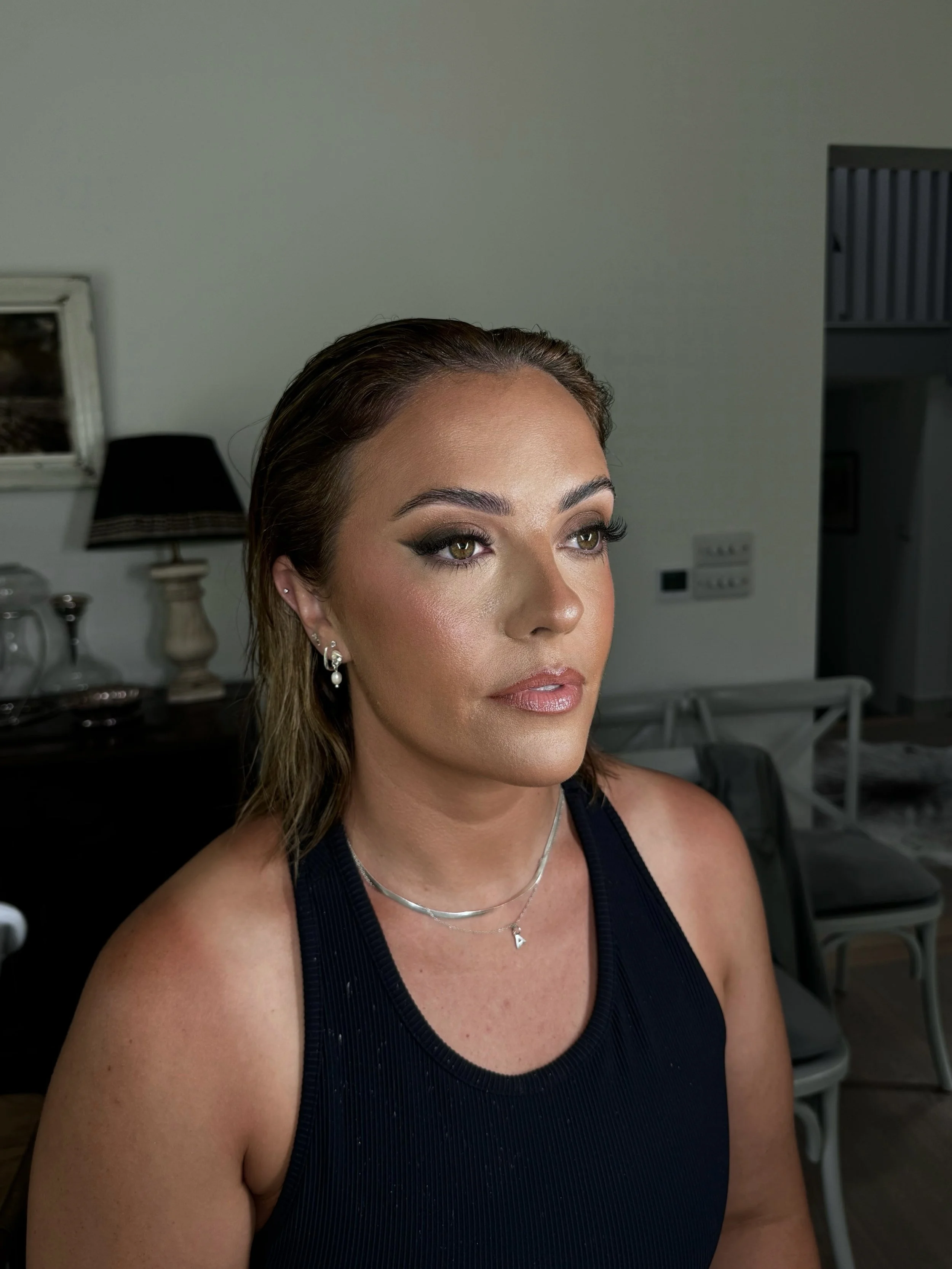 Close-up of a woman with makeup and jewelry, wearing a dark sleeveless top, sitting indoors with a neutral background.