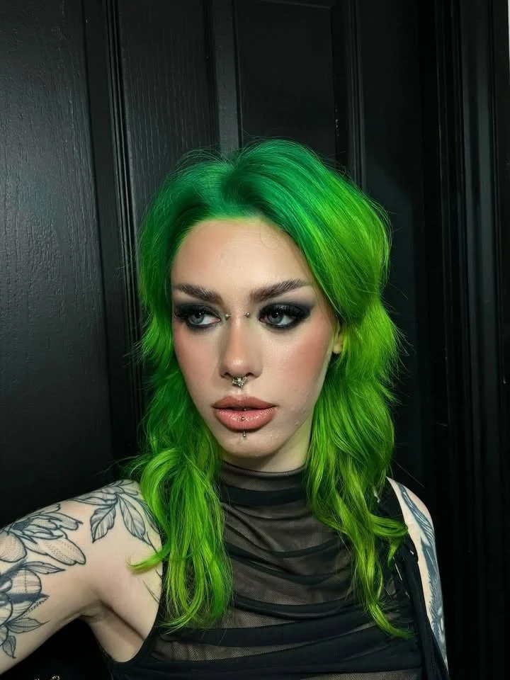 Woman with vibrant green hair, dark makeup, and tattoos on her arm, posing against a black wooden background.