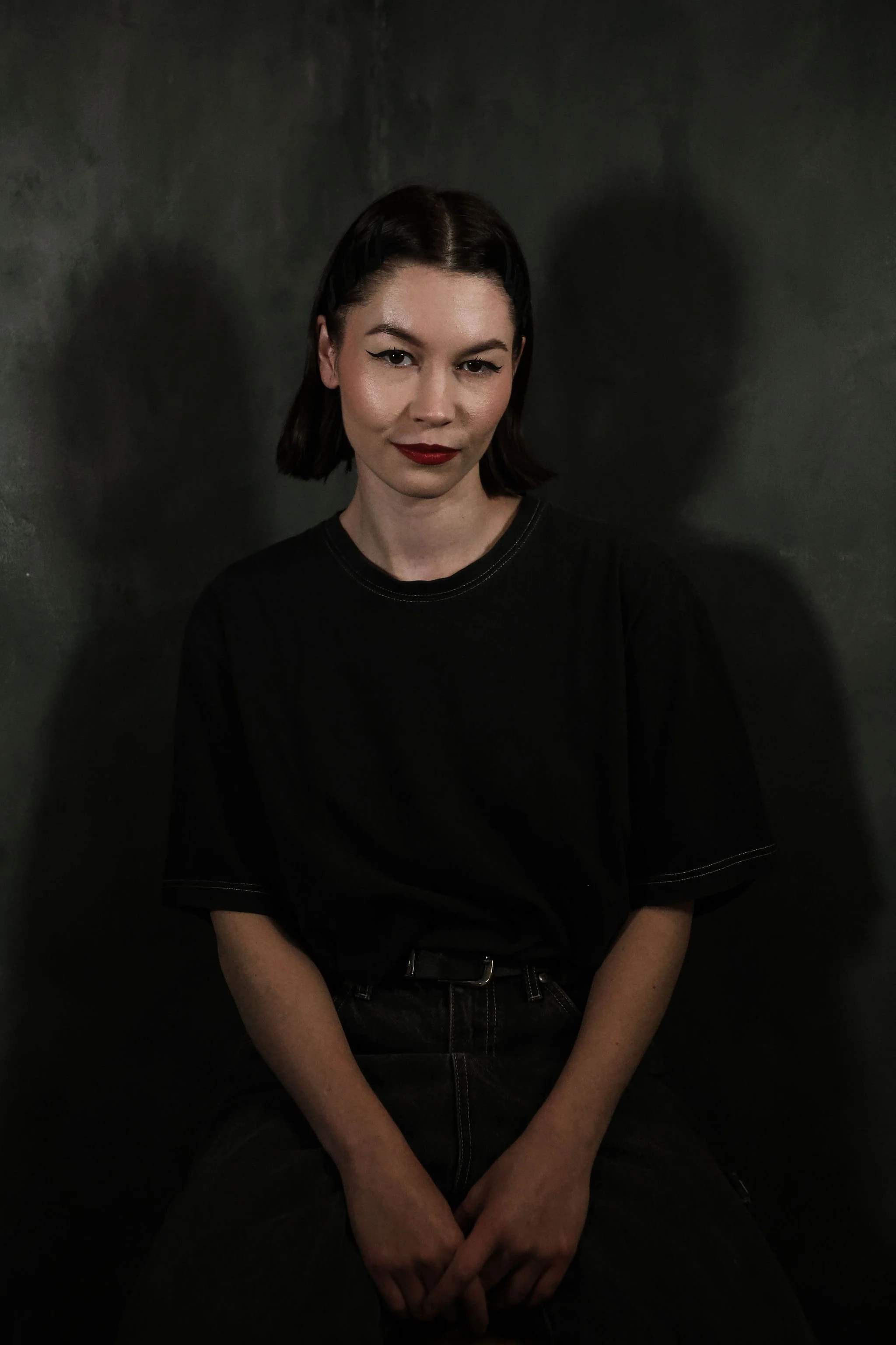 A young woman with dark hair, wearing a black t-shirt, sitting against a dark textured background, with a subtle confident expression.