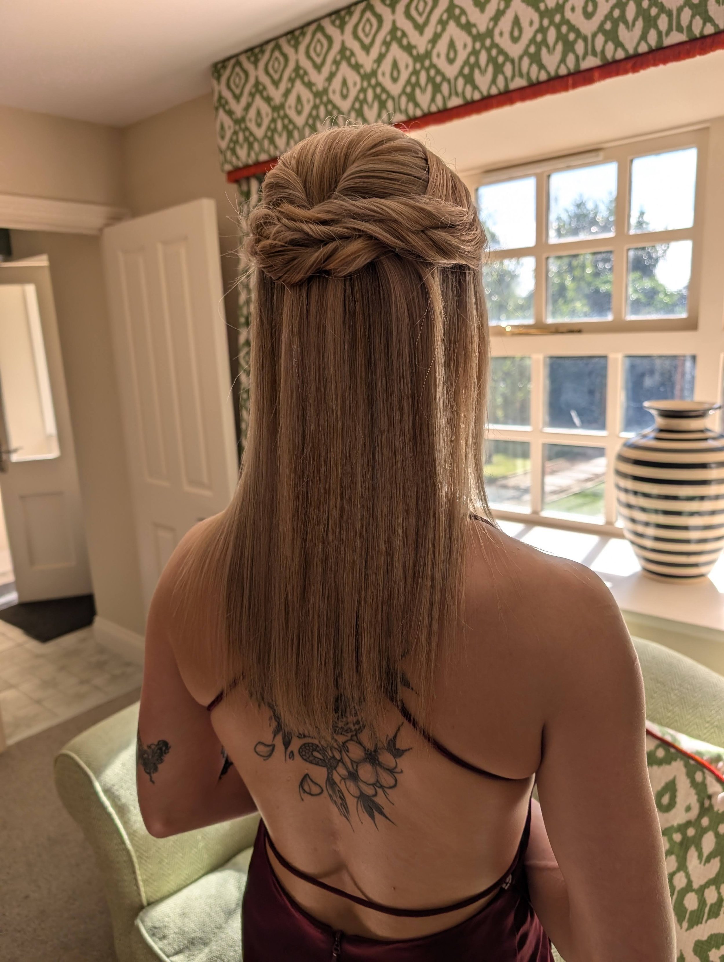 Rear view of a woman with long, straight blonde hair styled with a twisted braided crown on top, wearing a black strap dress, sitting on a light green chair in a room with a window, patterned wallpaper, and a striped vase on a white table.