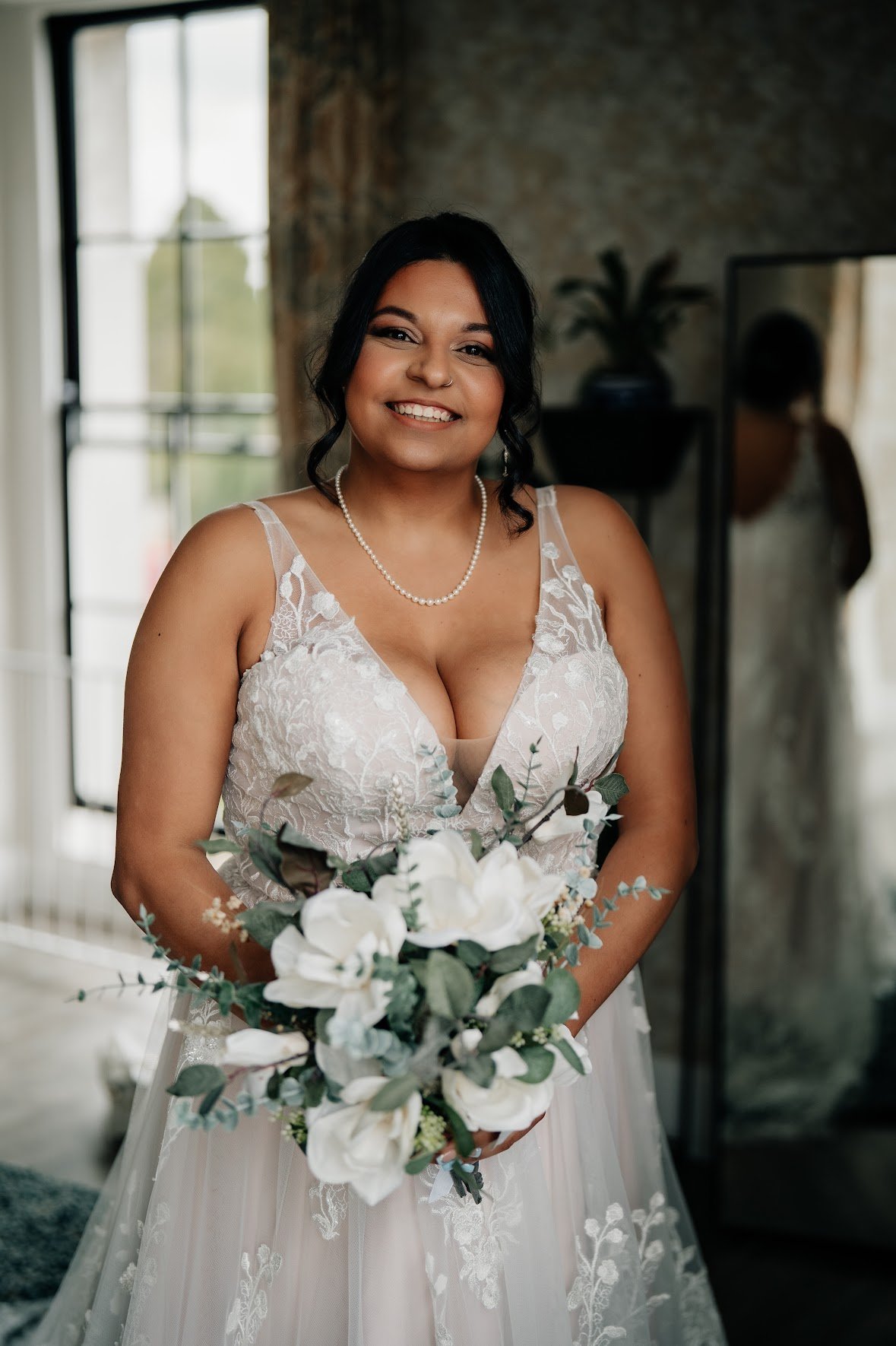 A smiling woman in a wedding dress holding a bouquet of white flowers, standing indoors with a window and mirror in the background.