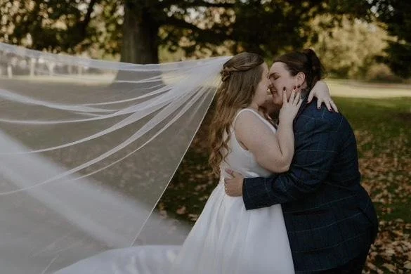 Two women embrace and kiss outdoors during a wedding, with one wearing a white wedding dress and a veil, and the other in a dark suit, surrounded by trees and grass.