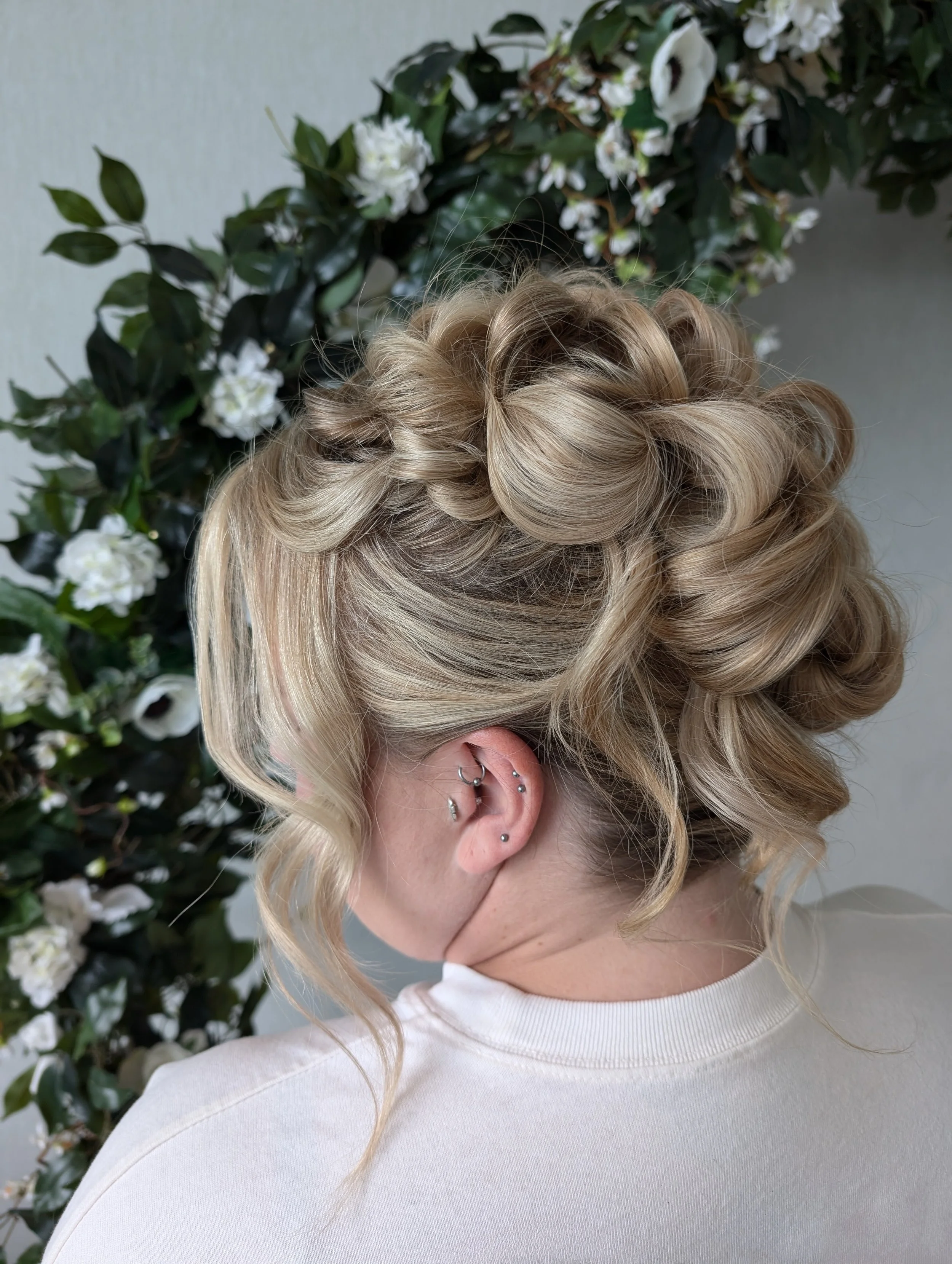 A woman with blonde, curled hair and multiple piercings on her ear, looking down with her face partially covered, standing in front of a floral background.