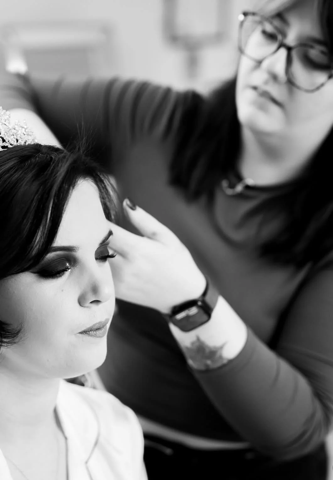 A makeup artist applies makeup to a woman with closed eyes, in a black and white photo.