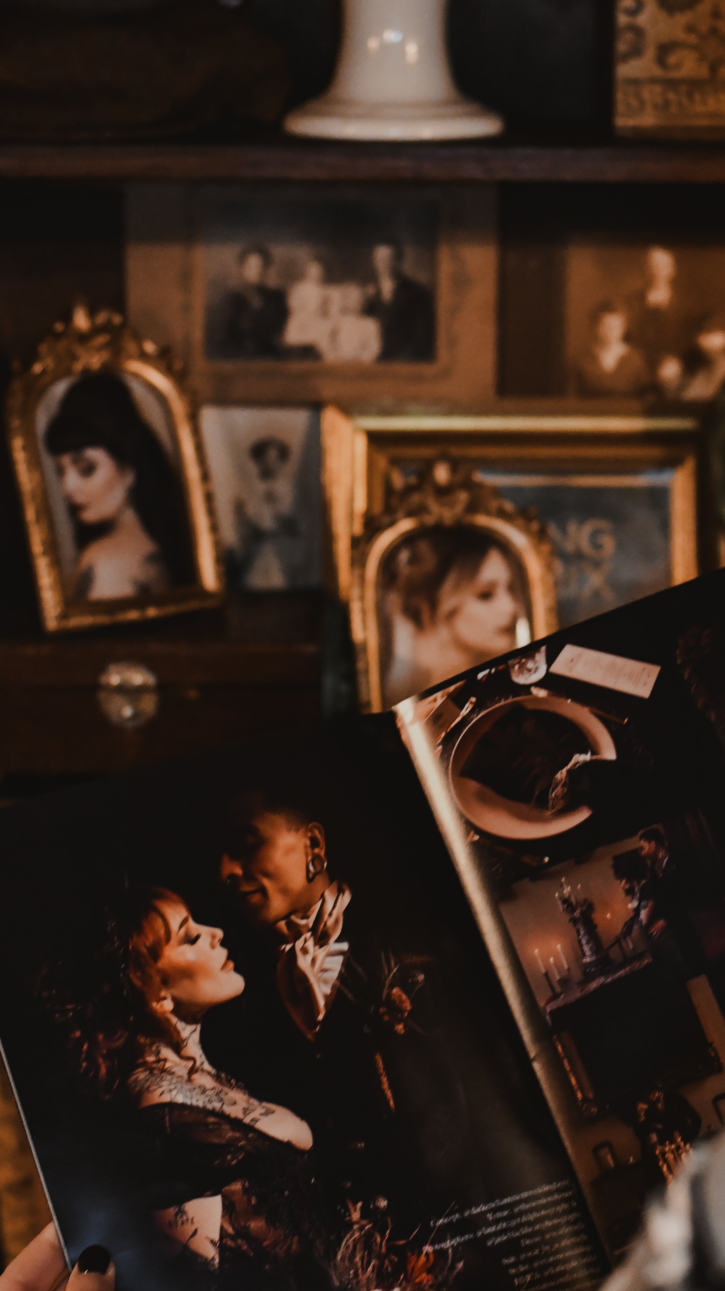 A person holding an open magazine with a photo of a woman and a man in formal attire, in lead of a background filled with vintage framed photographs of women and children on a wooden wall and shelf.