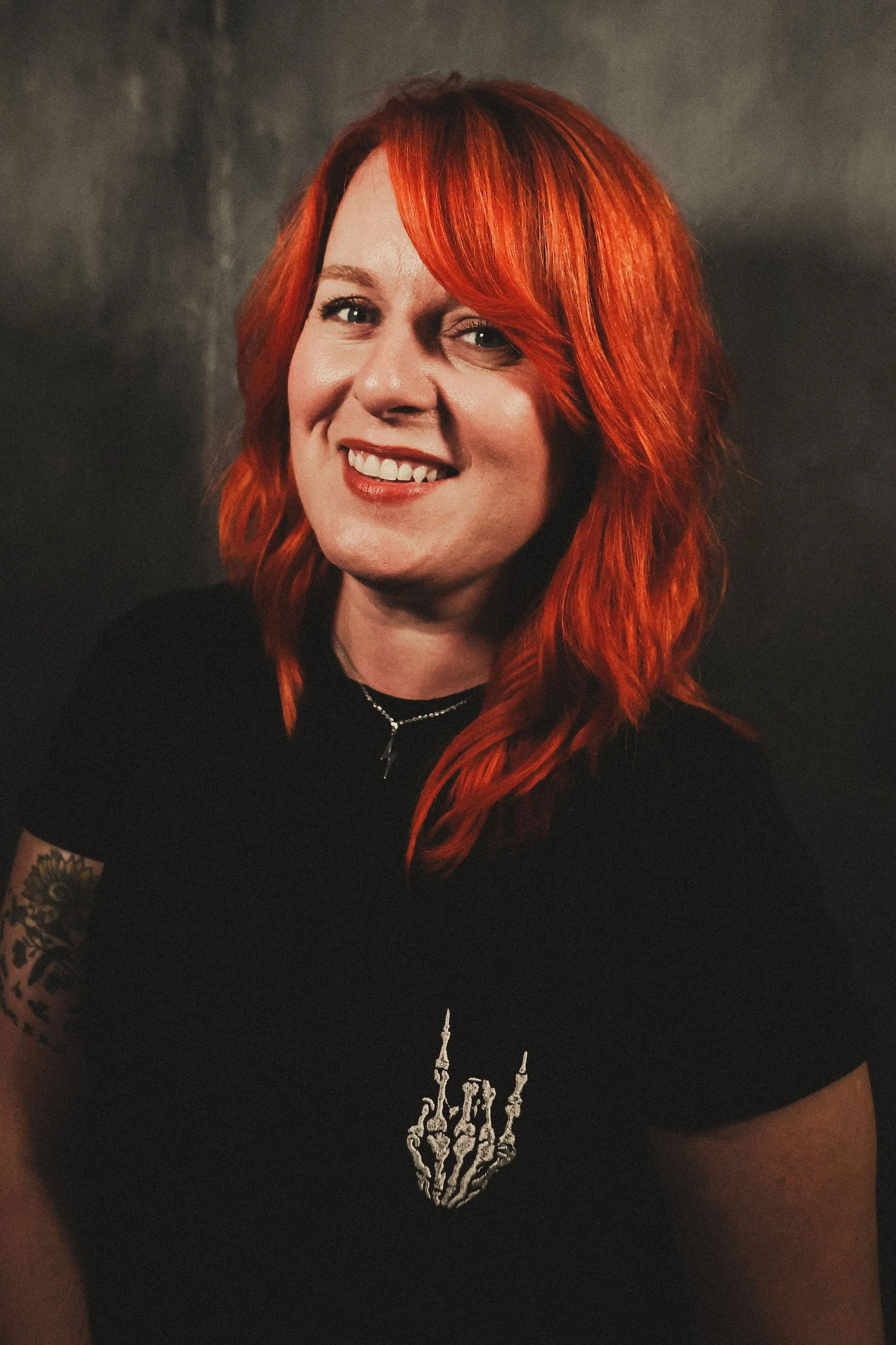 A woman with bright red hair smiling and wearing a black t-shirt with a white skeletal hand making a rock on gesture.