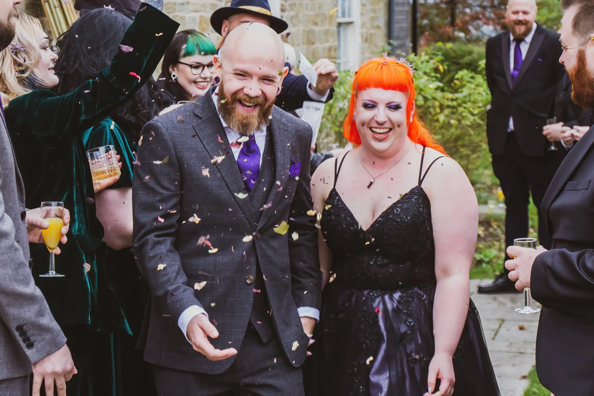 People celebrating outdoors, with a man and a woman smiling at the center, surrounded by others holding drinks and throwing confetti, during a festive event.