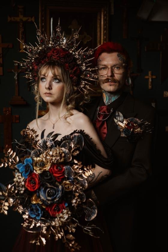 A woman with a floral crown and bouquet, and a man with glasses and makeup, standing together in a dark, ornate room with crosses on the wall.