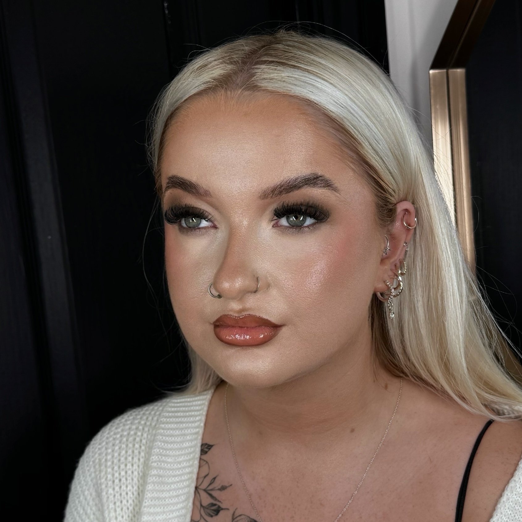 Close-up of a woman with platinum blonde hair, green eyes, and multiple earrings, wearing makeup, a nose ring, and a white sweater with a tattoo on her chest.