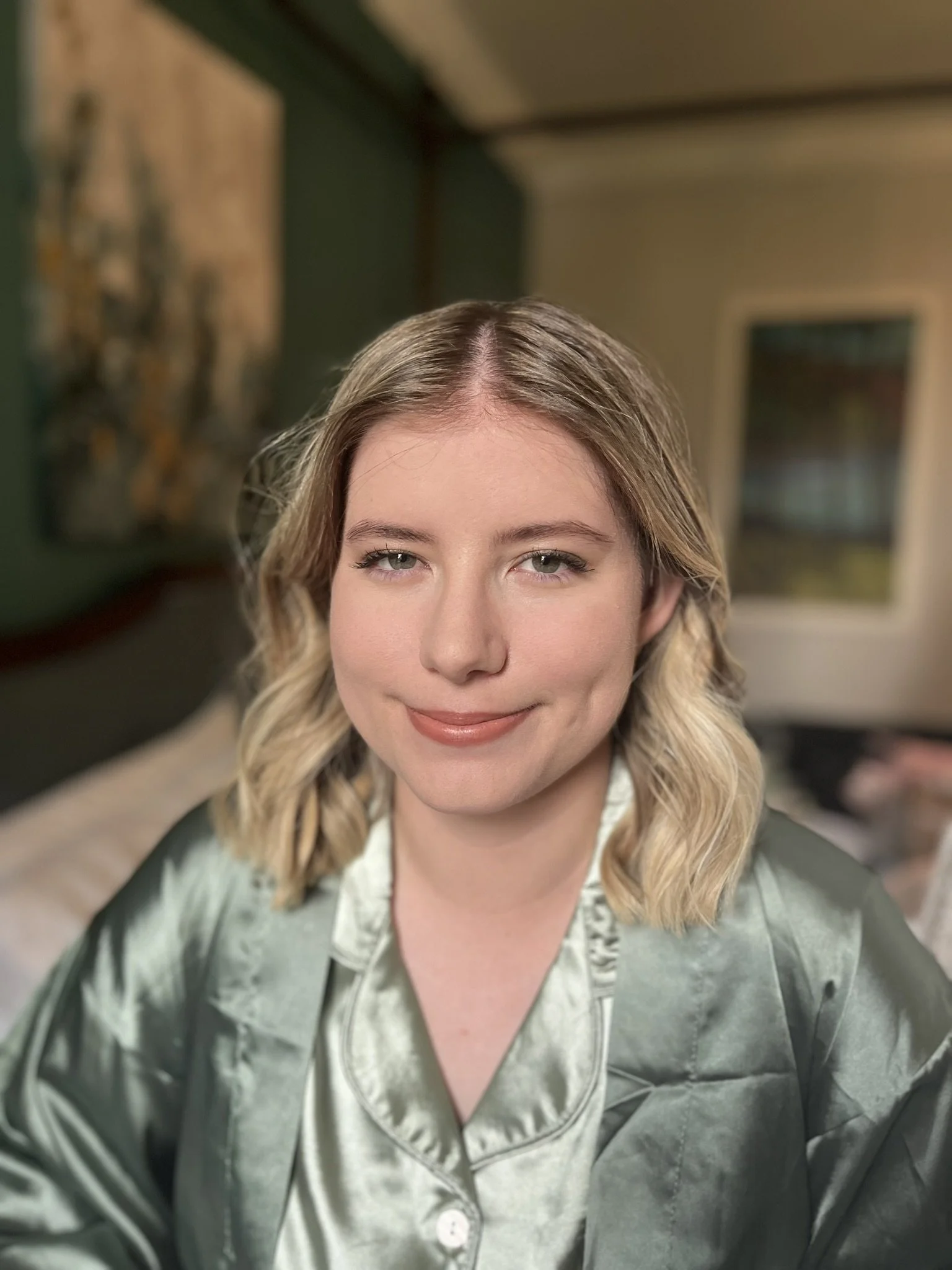 A woman with light blonde wavy hair, wearing a satin olive green pajama top, smiling in a bedroom with a bed, window, and artwork on the green wall.
